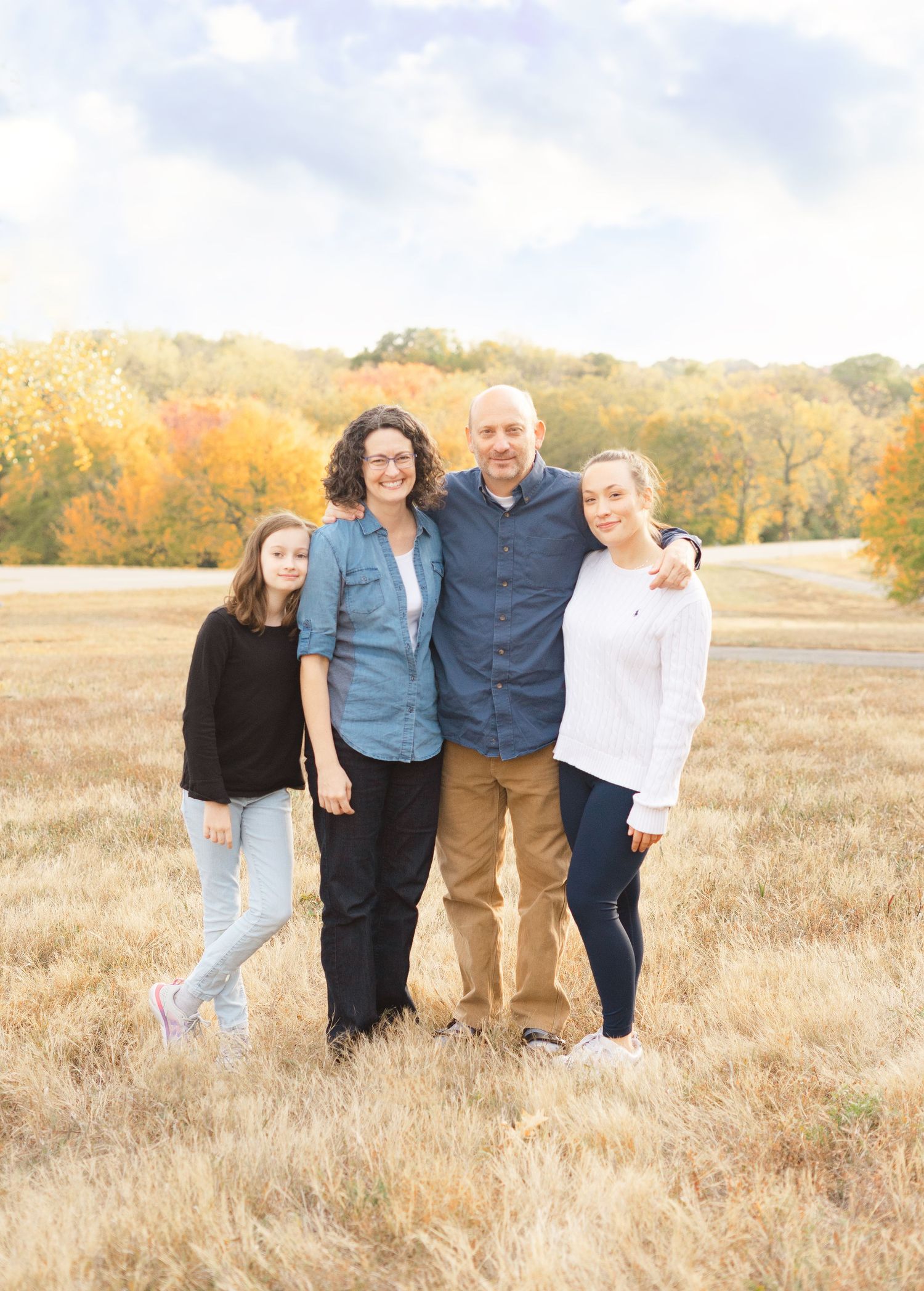 A group of four people pose together in a fall field with autumn foliage visible in the background.