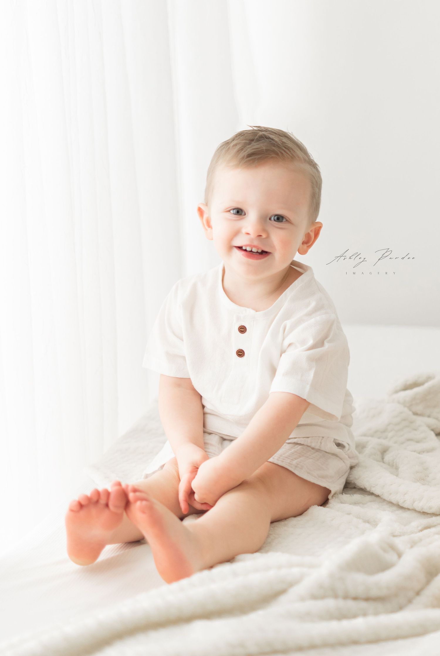 Small child wearing white henley shirt and khaki shorts sits on white bedding with a bright happy expression.
