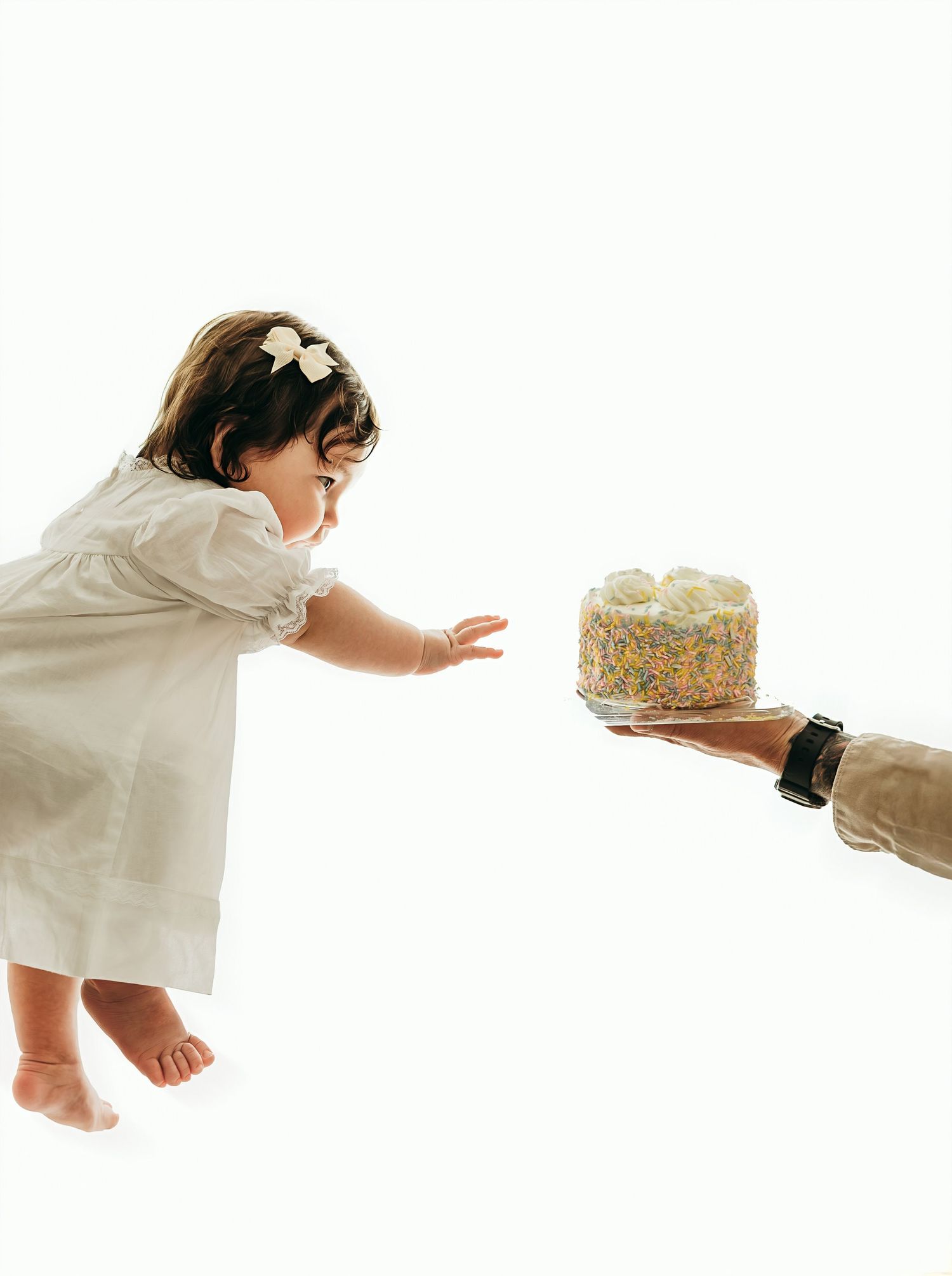A baby girl in a white dress reaches eagerly toward a colorful sprinkle cake held by an adult hand.