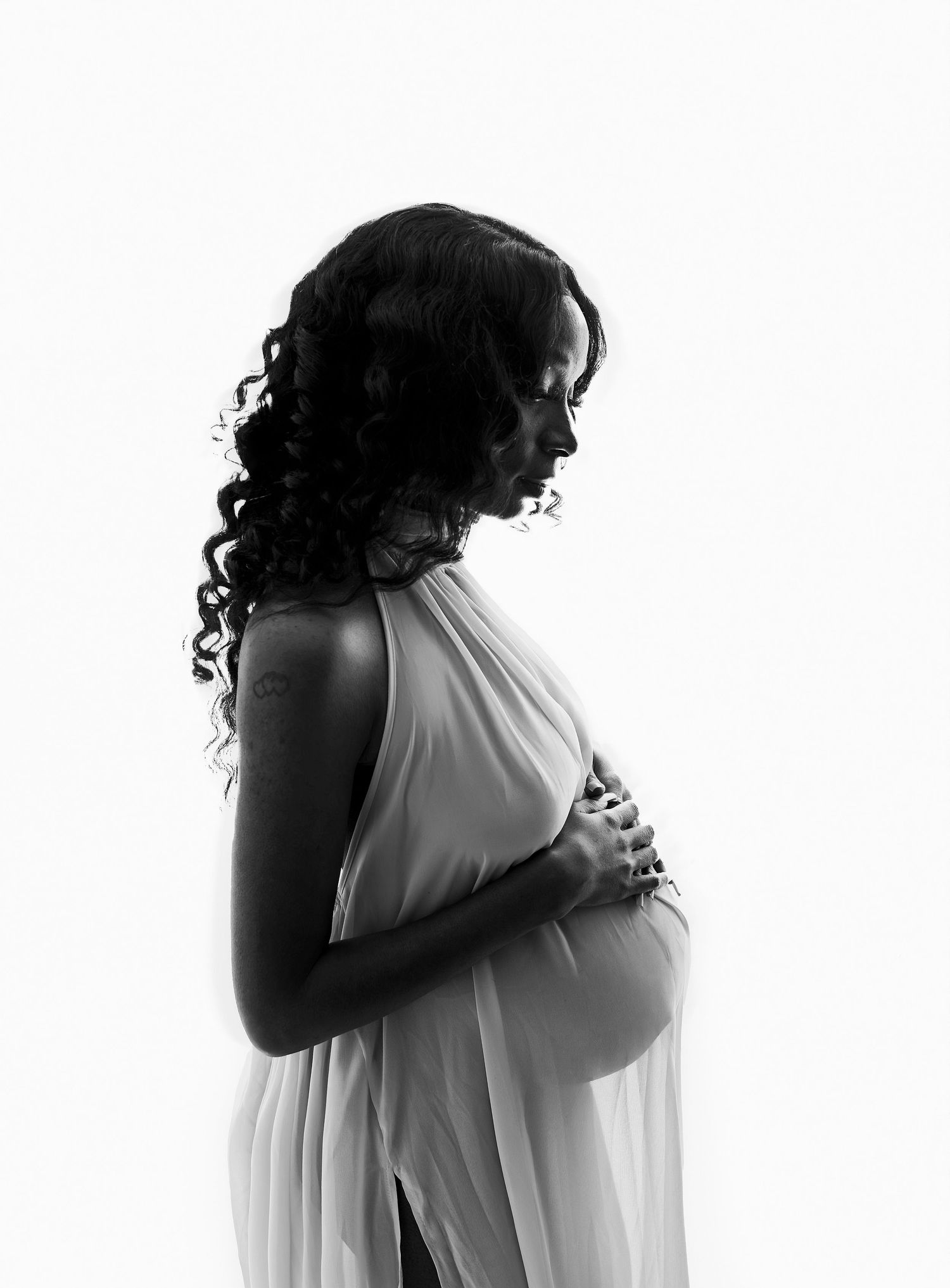 Black and white maternity photo of a pregnant woman in sheer draped fabric, cradling her baby bump in profile view.