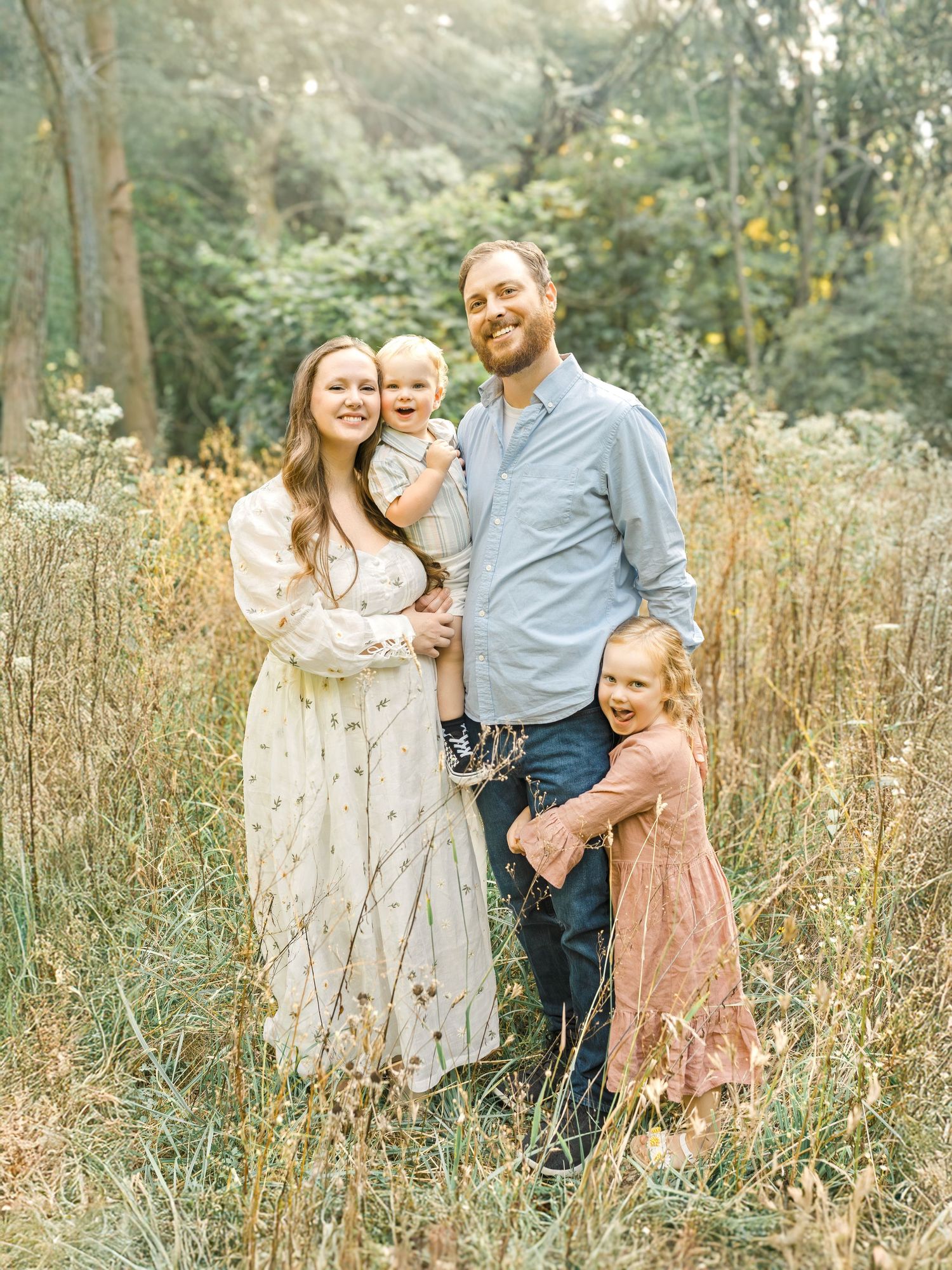 A smiling family of four standing close together during a soft, natural outdoor session in Kansas City.