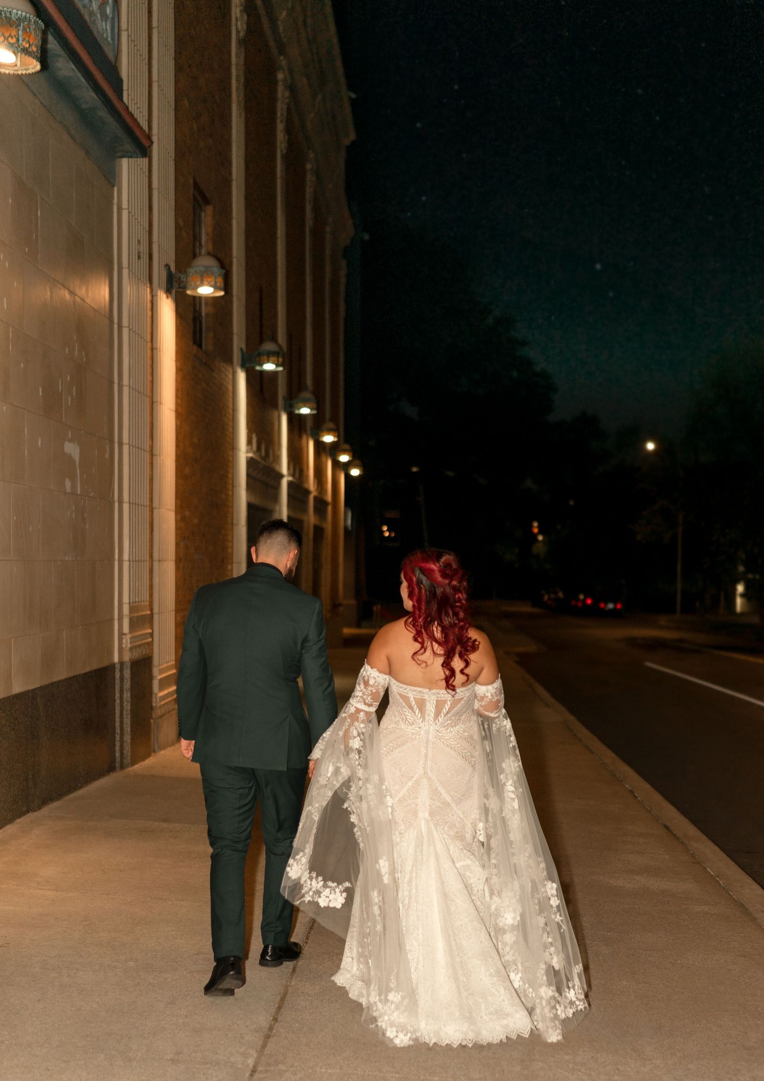 Couple walking down an illuminated sidewalk at night, with a bride in a white lace dress with flowing cape sleeves.