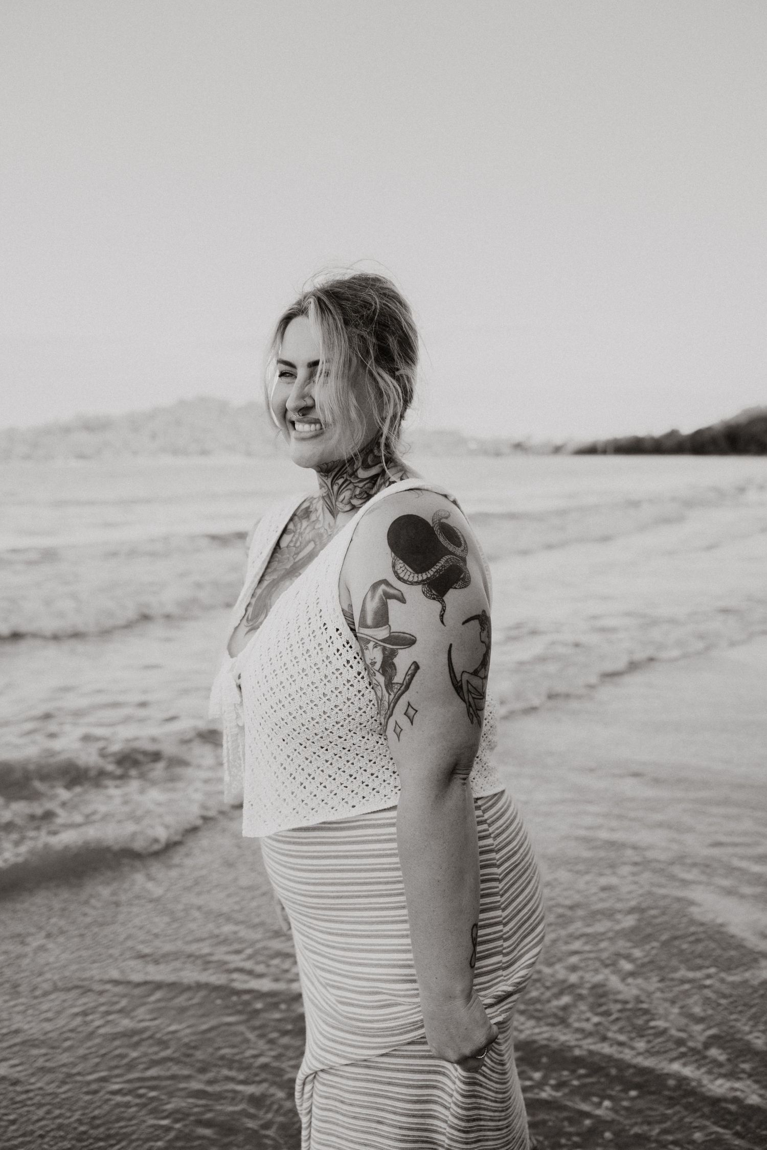 Tattooed woman in white outfit smiling on a beach in black and white photo.