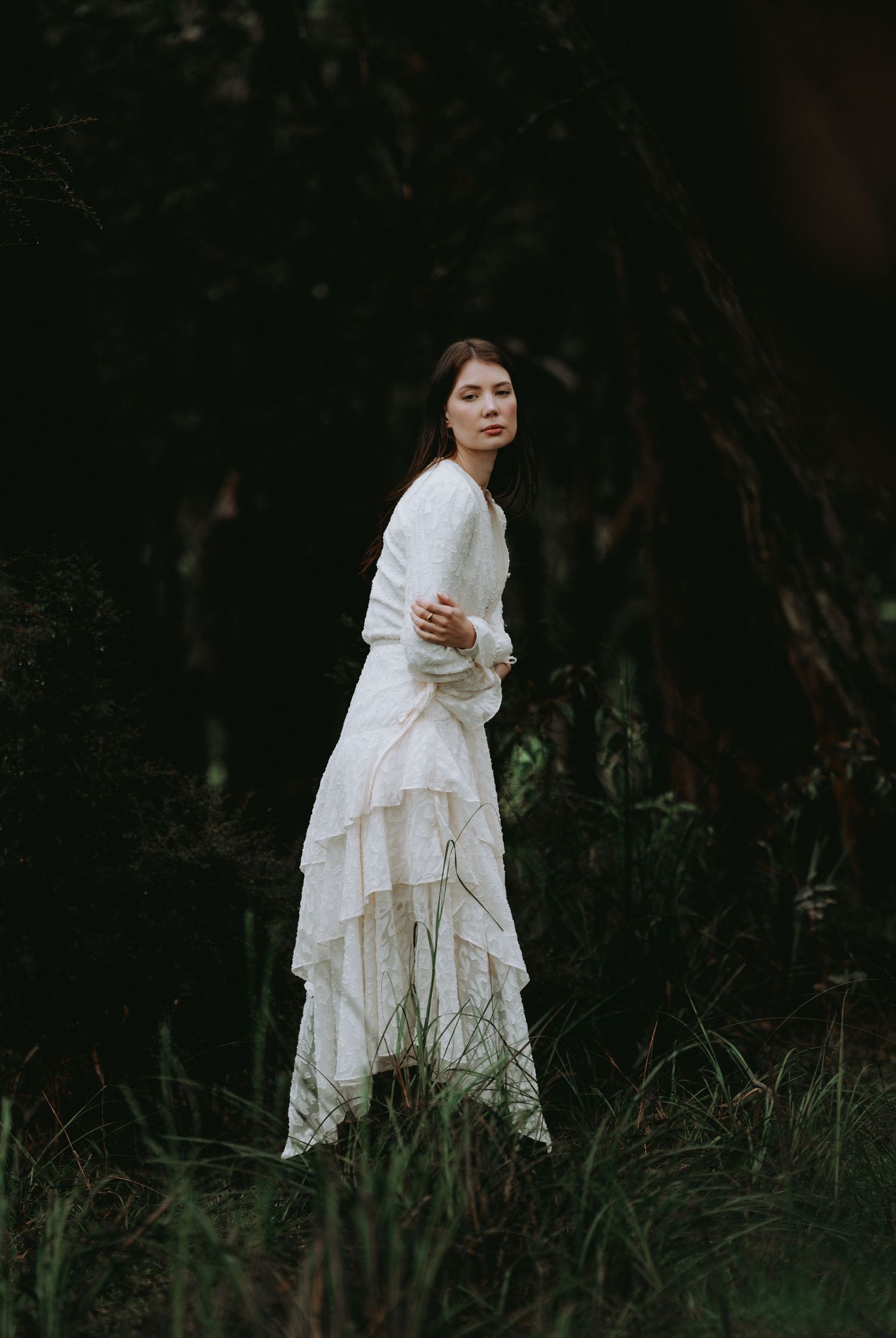 Brunette woman in elegant white tiered lace dress standing in a dark moody forest setting.