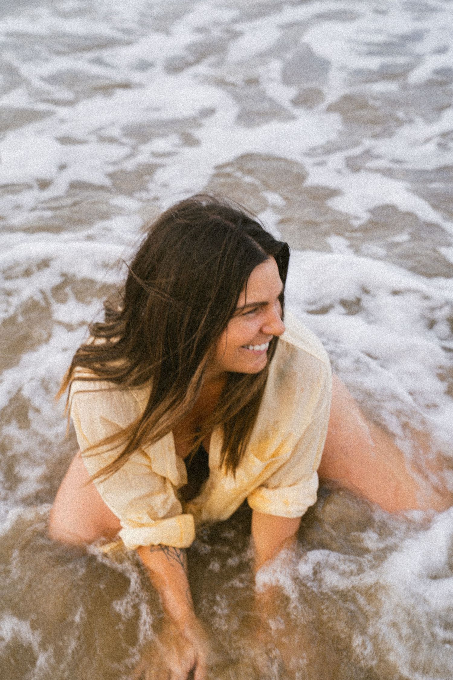 Smiling brunette woman in yellow shirt playing in ocean waves on a sandy beach.