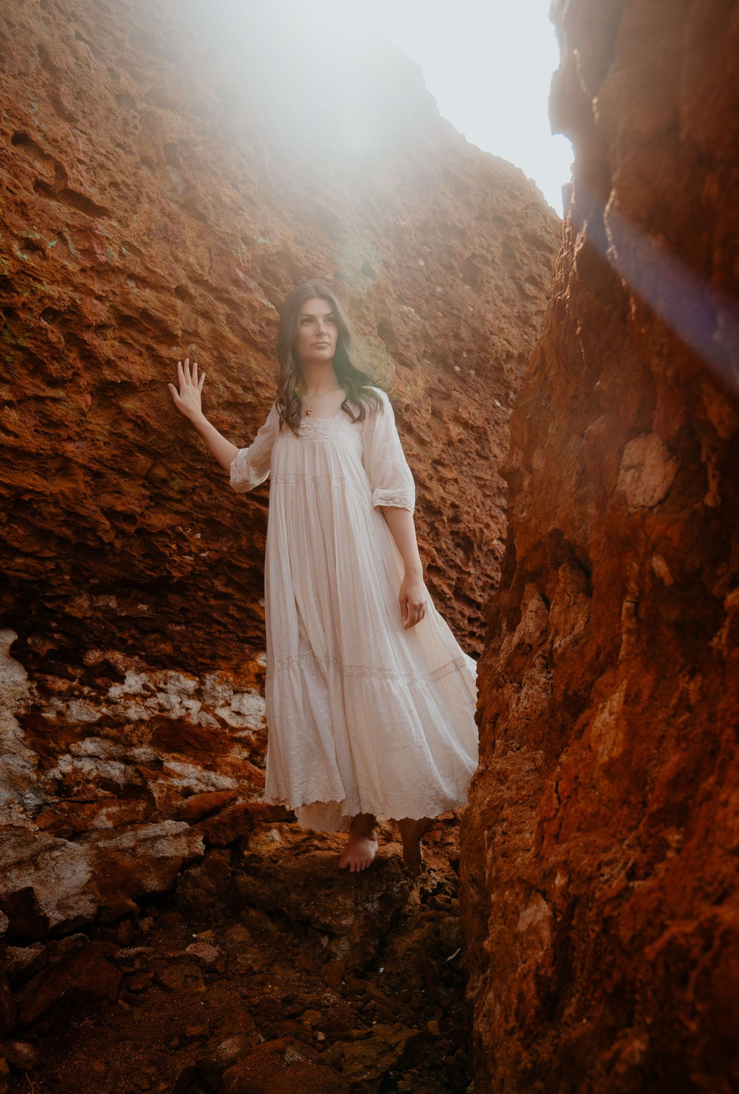 Woman in flowing white dress stands between red rock canyon walls in golden sunlight.