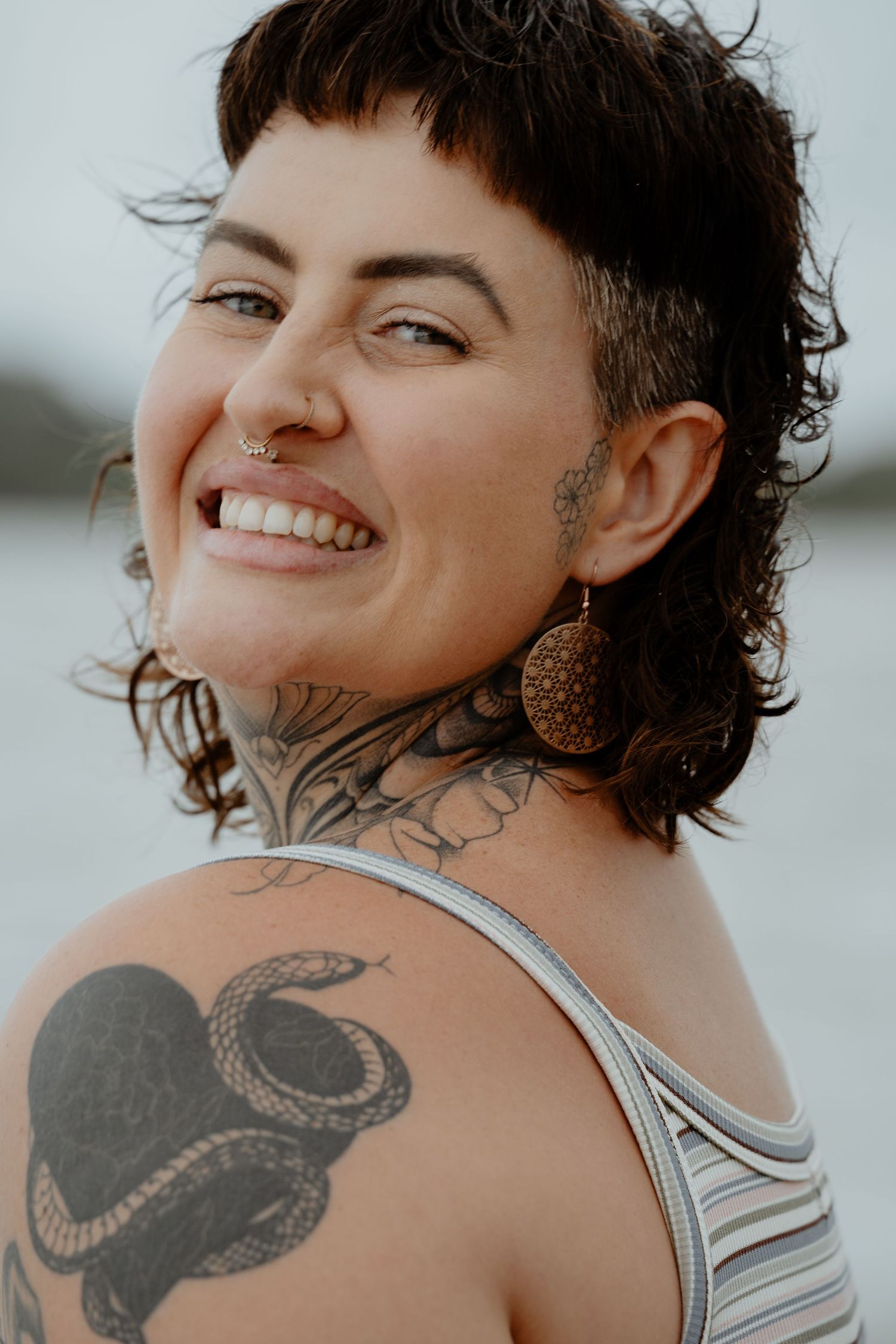 Smiling tattooed woman with shaved sides and nose ring wearing striped tank top near water outdoors.