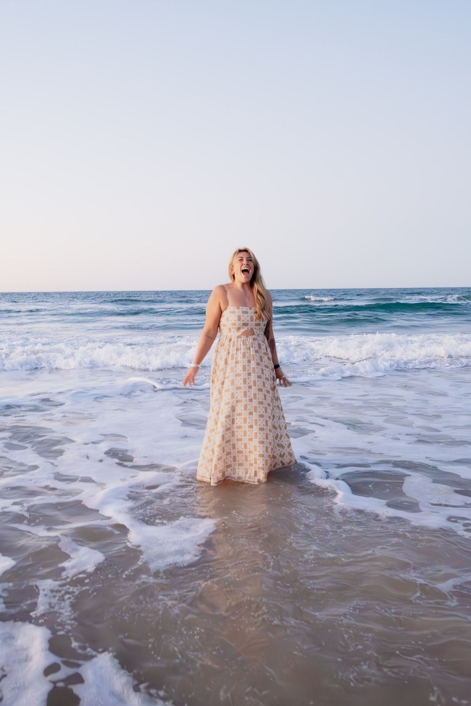 Blonde woman in a floral maxi dress reacting with surprise as ocean waves wash over her feet.