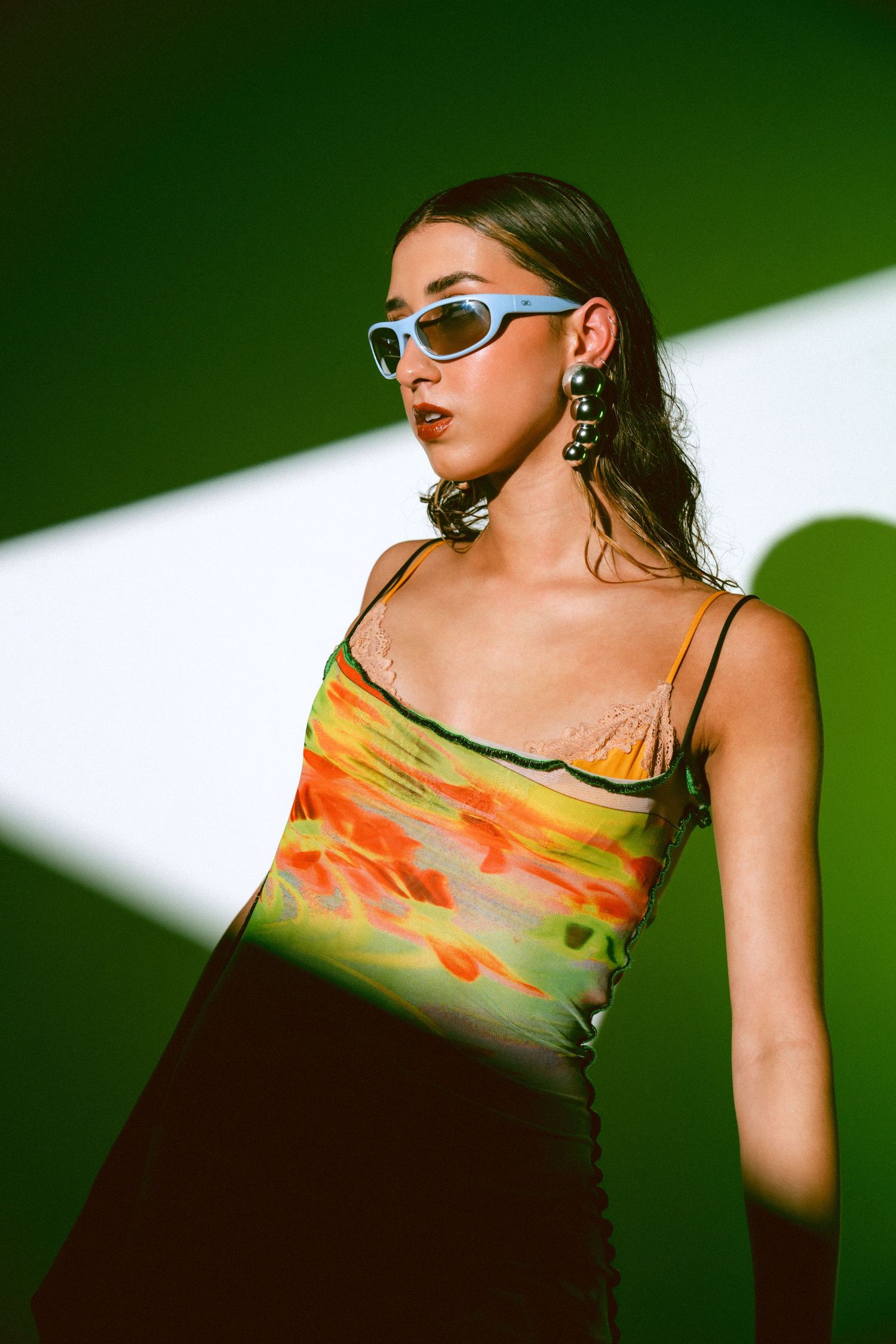 Young woman in colorful tie-dye slip dress and blue sunglasses posing against green background.