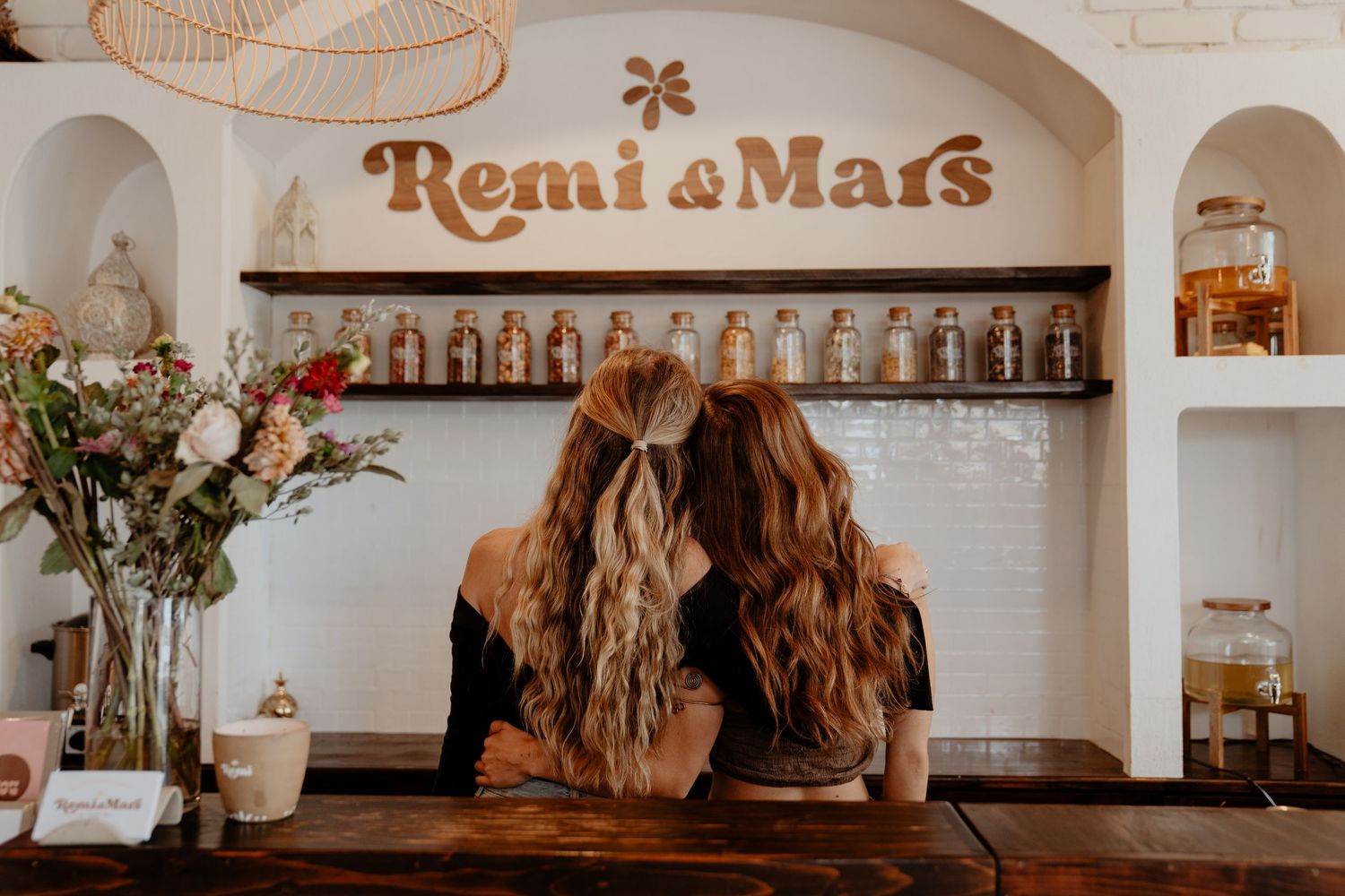 Two women with long hair sitting at wooden counter beneath Remi and Mars sign with glass jar display.