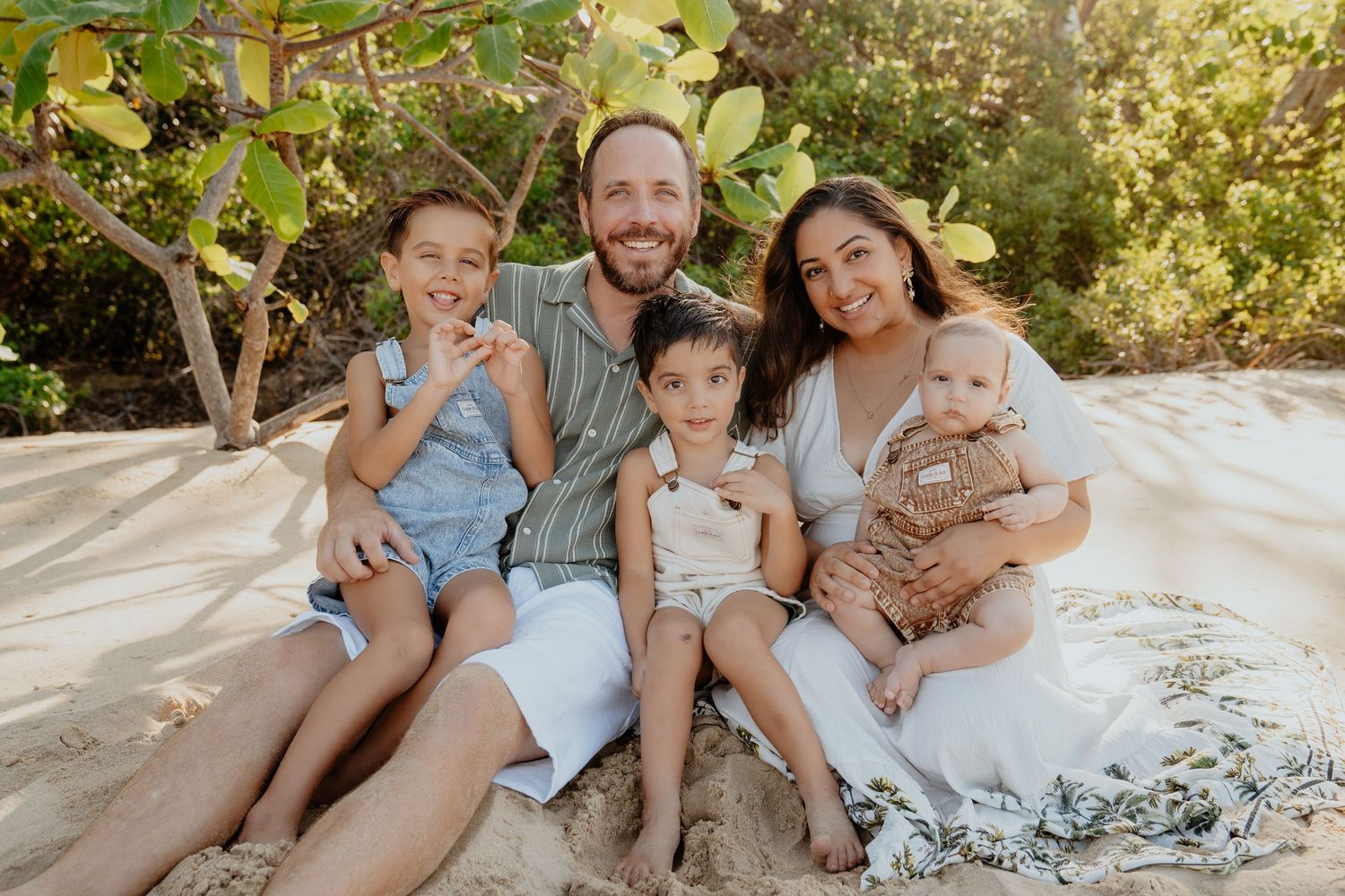 Happy family of five sits together on sandy tropical beach in warm sunlight.