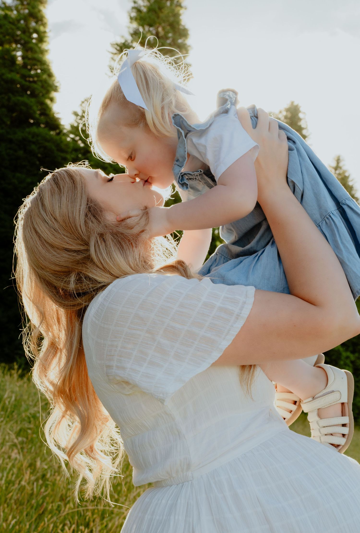 Pregnant mother in white dress lifting and kissing her blonde toddler daughter outdoors.