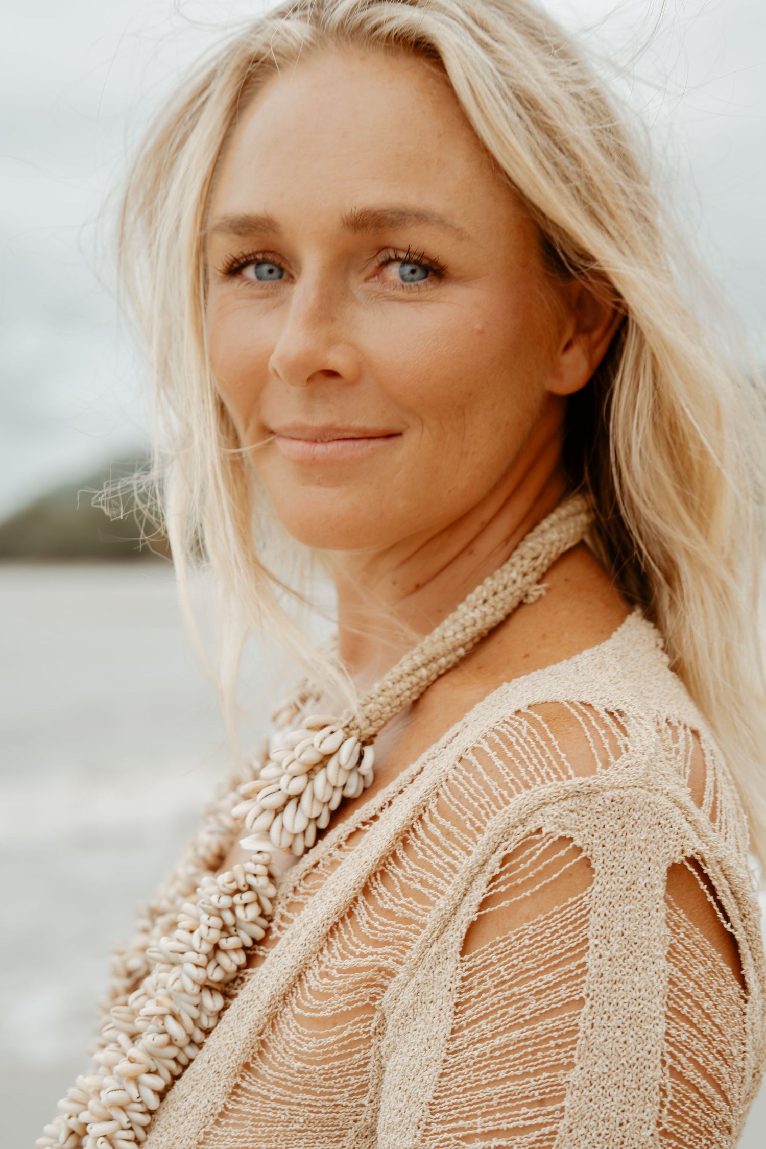 Blonde woman with blue eyes wearing a shell necklace and beige top at the beach.