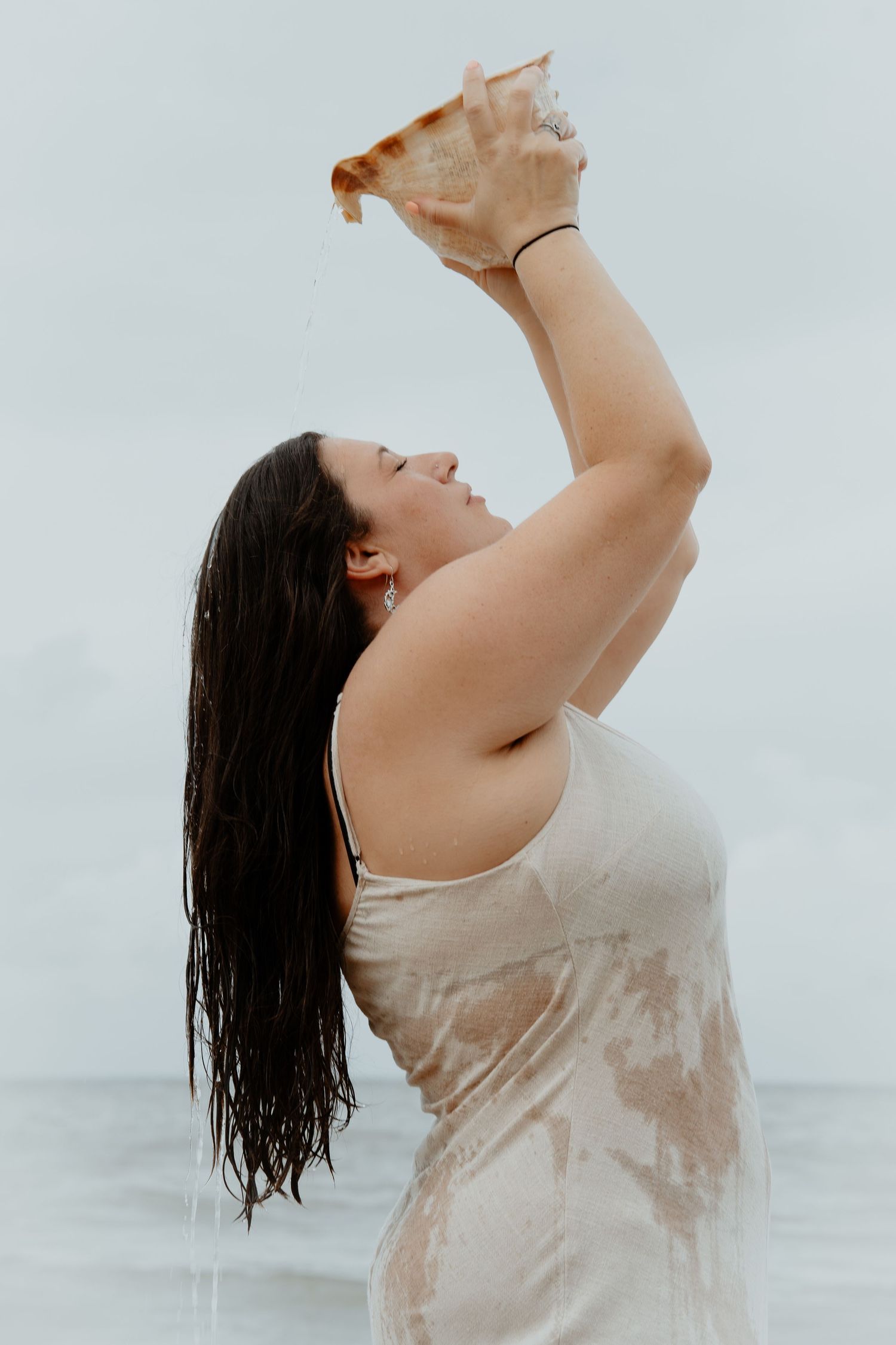 Woman in wet beige dress lifting a conch shell overhead while standing in the ocean outdoors.