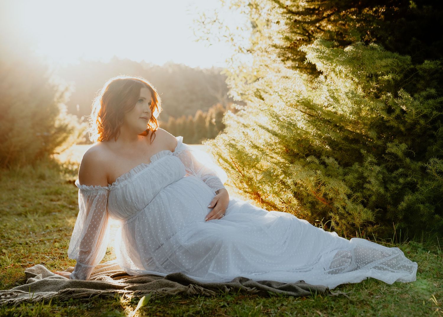 A pregnant woman in a flowing white off-shoulder gown reclines gracefully on grass in golden sunset light outdoors.