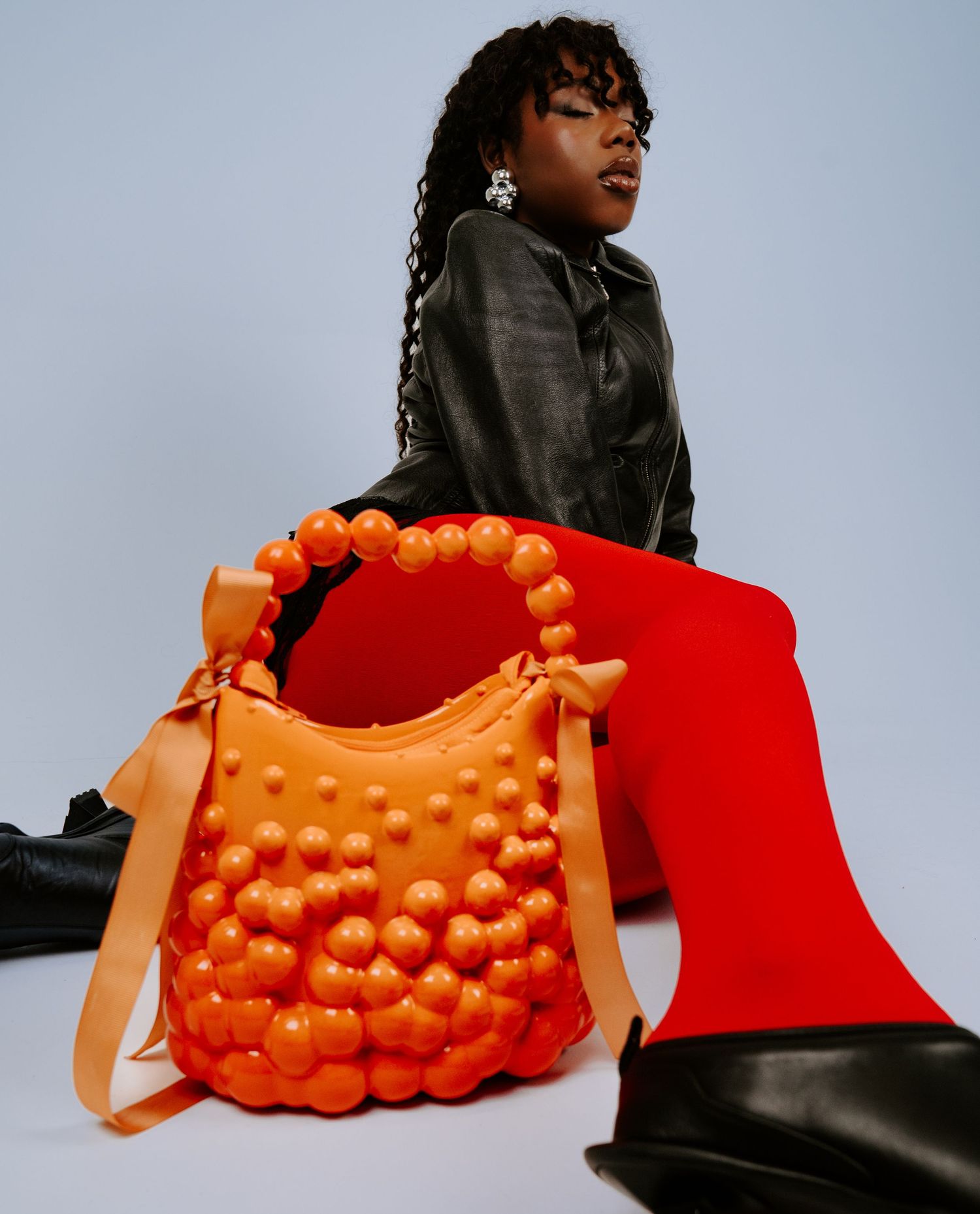 Model in black leather jacket posing with orange bubble-textured handbag and red trousers.