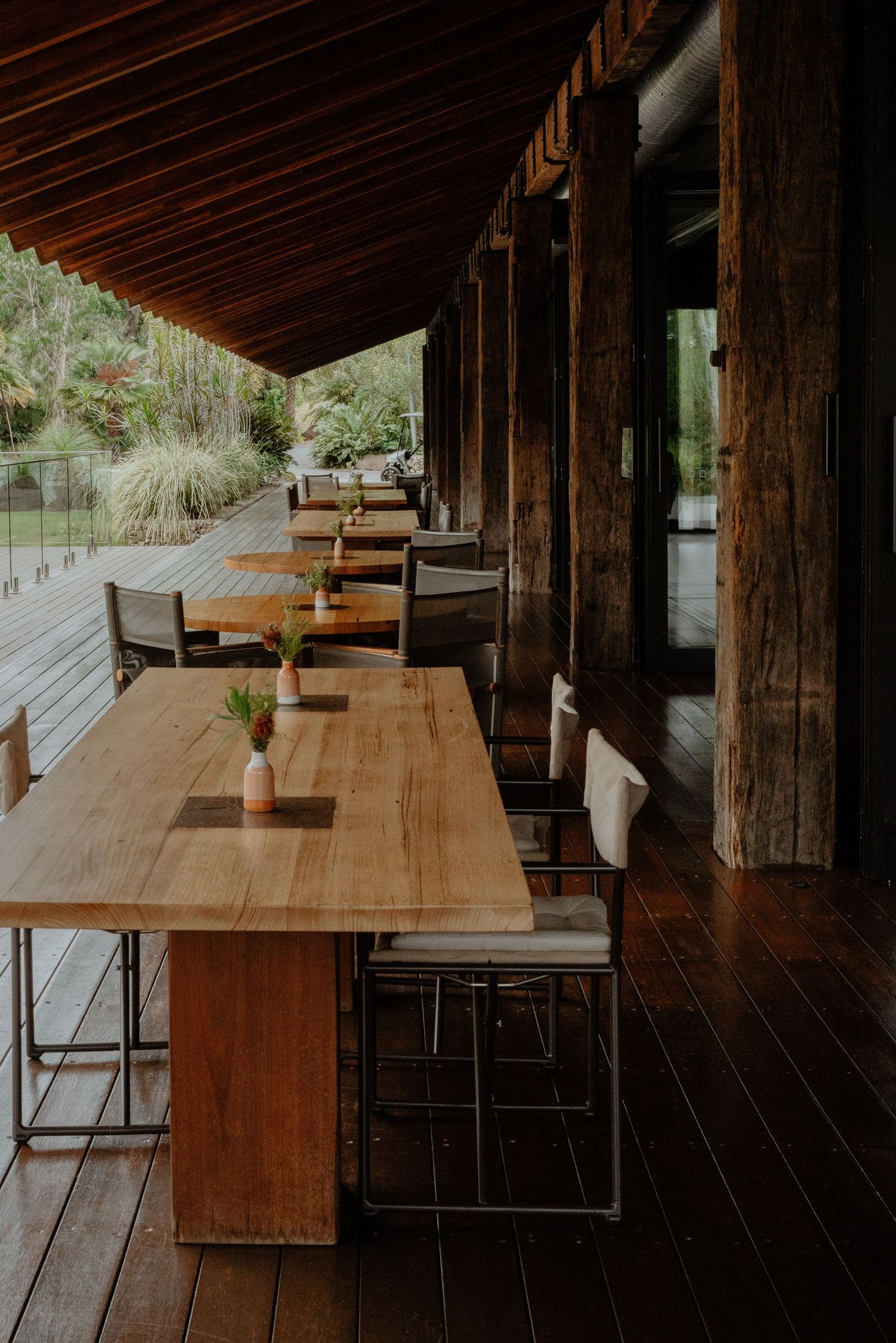 Wooden outdoor dining tables and chairs on a covered deck surrounded by lush greenery.