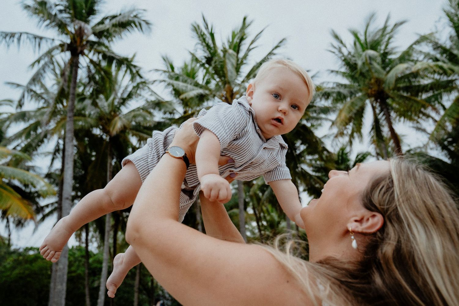 Mother joyfully lifting baby in the air surrounded by tropical palm trees outdoors.