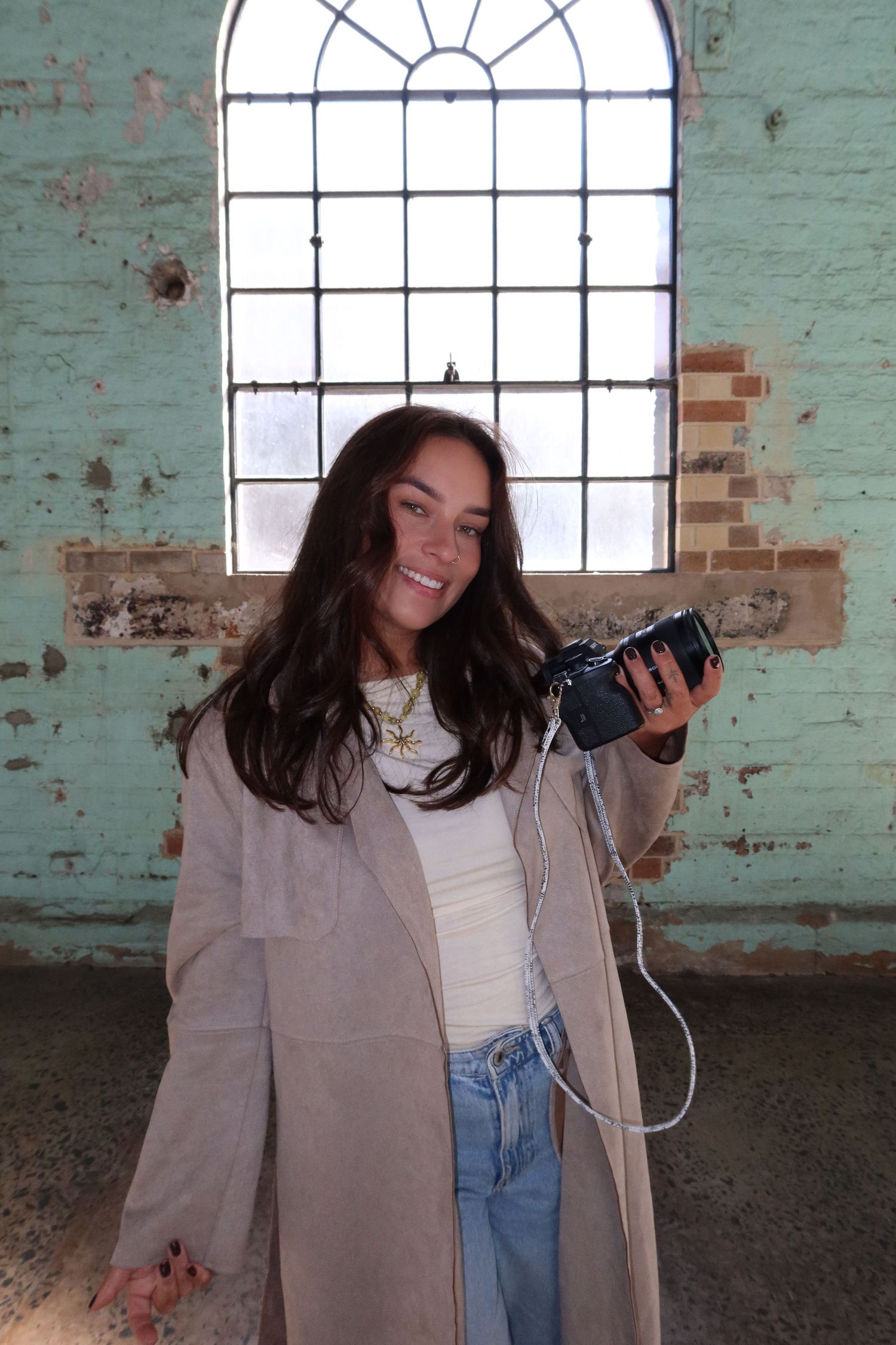 Smiling young woman holding vintage camera in rustic brick building with arched window.