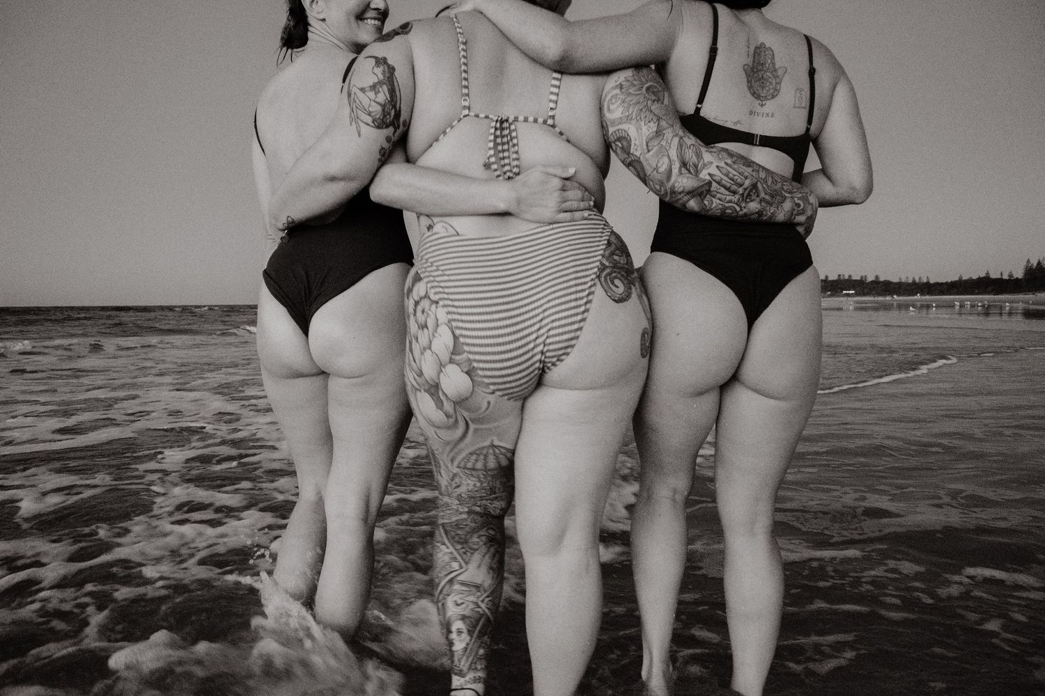 Black and white photo of four tattooed women in swimsuits hugging with backs to camera in ocean water.