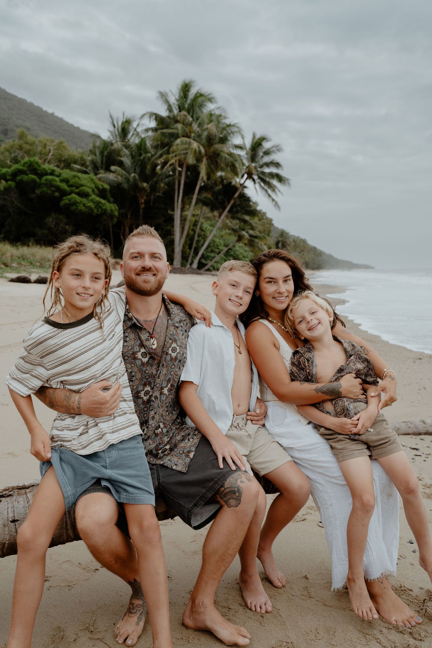 Smiling family of five sitting together on a log on a beautiful tropical sandy beach.