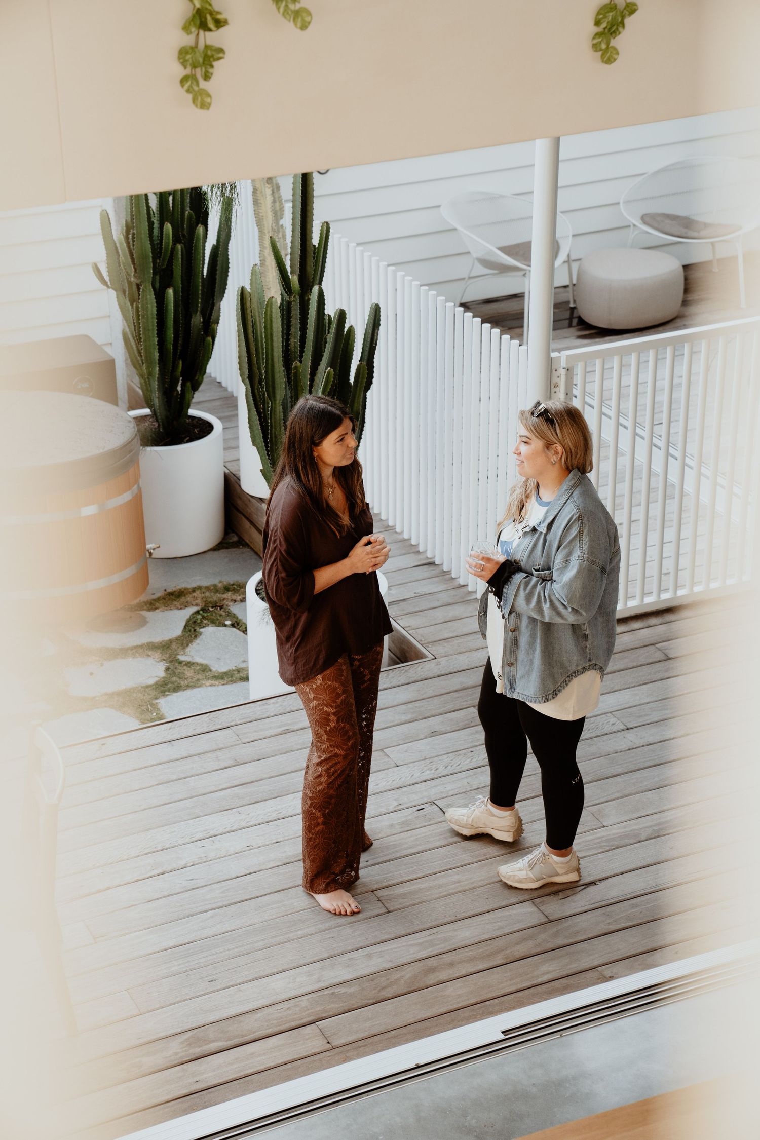 Two women having a conversation on a wooden deck surrounded by tall cactus plants in white pots.