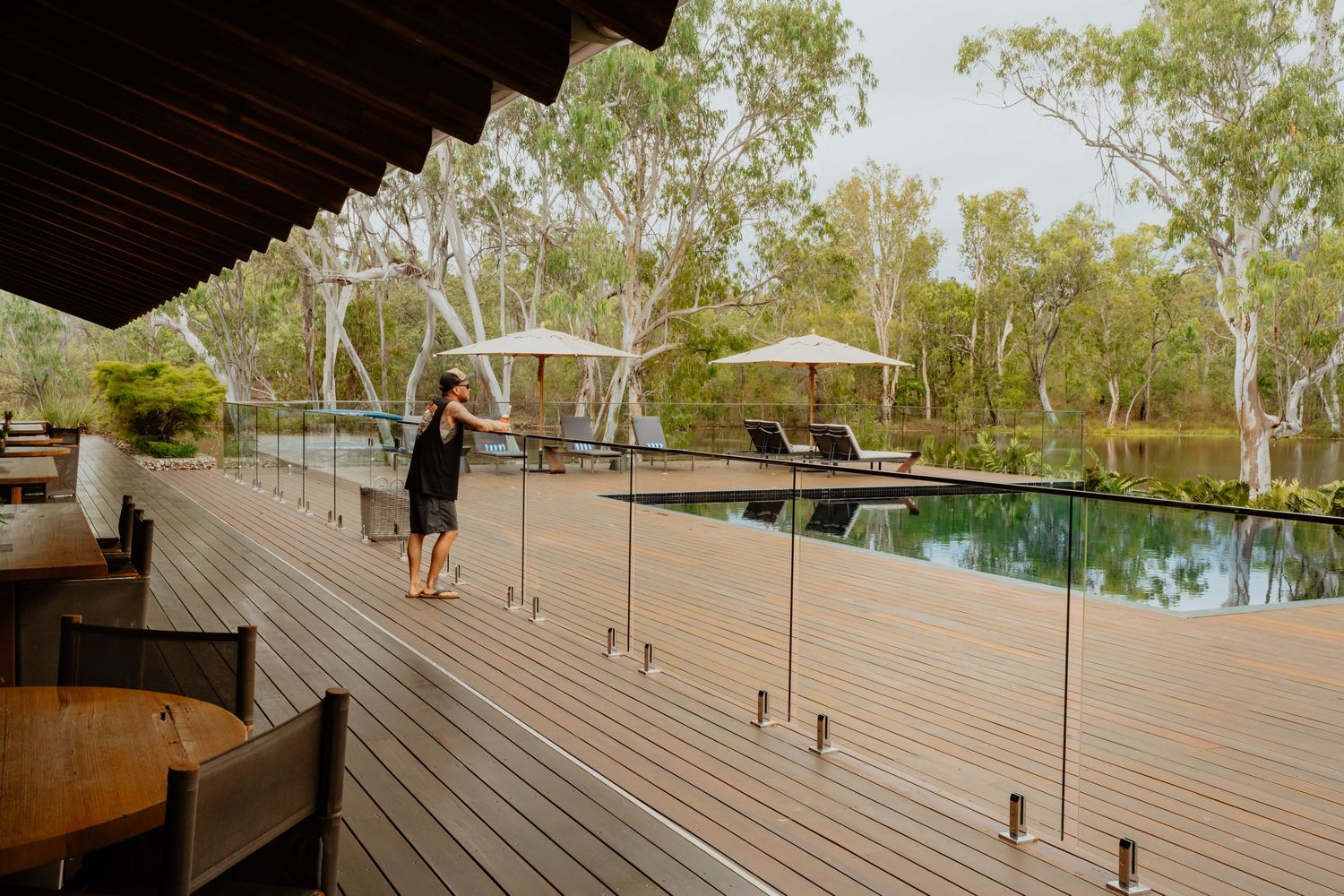 Woman standing on a wooden deck with glass railings overlooking a serene pool among eucalyptus trees.