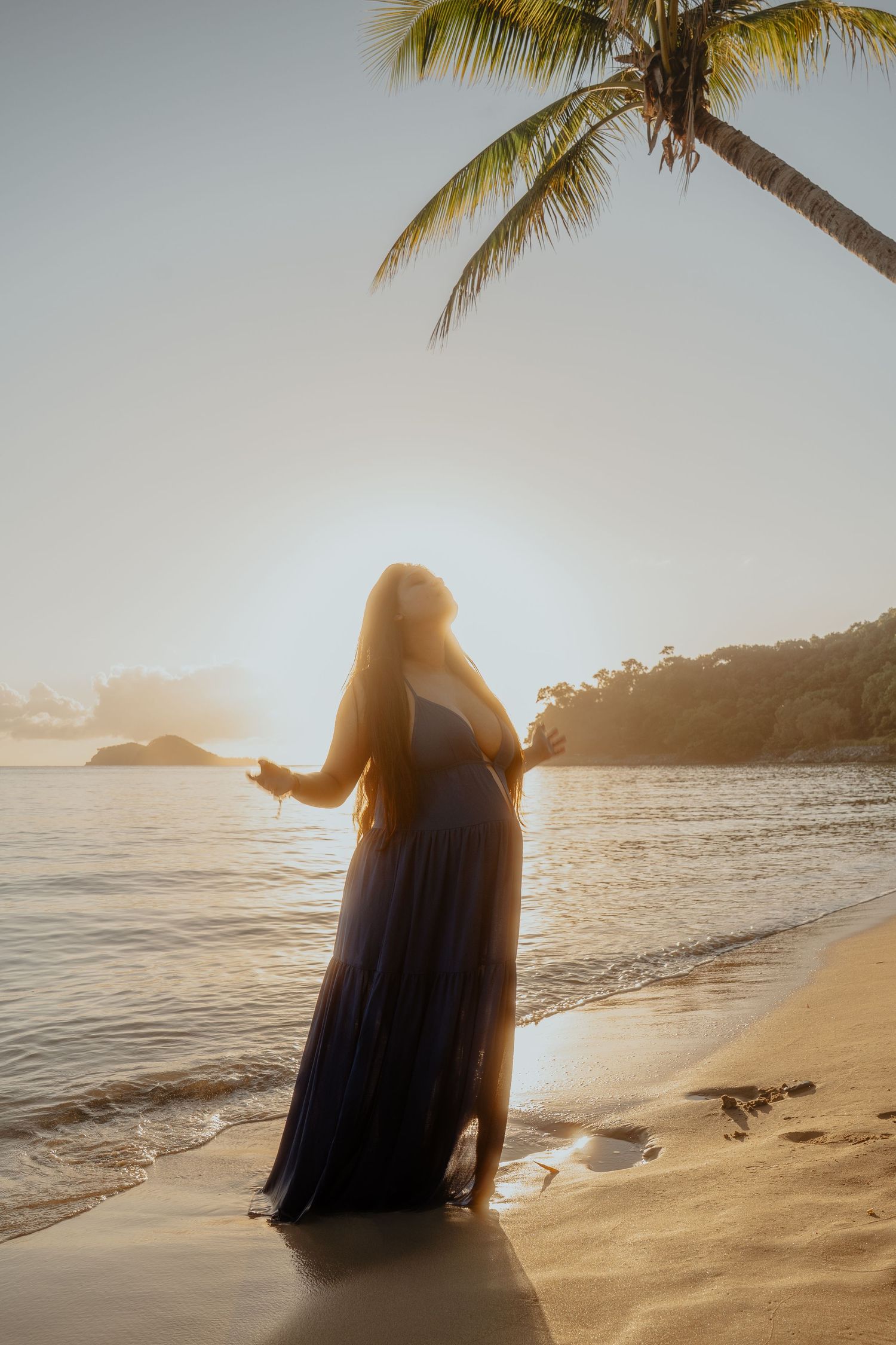 Silhouette of pregnant woman in flowing blue dress on sandy beach at golden sunset.