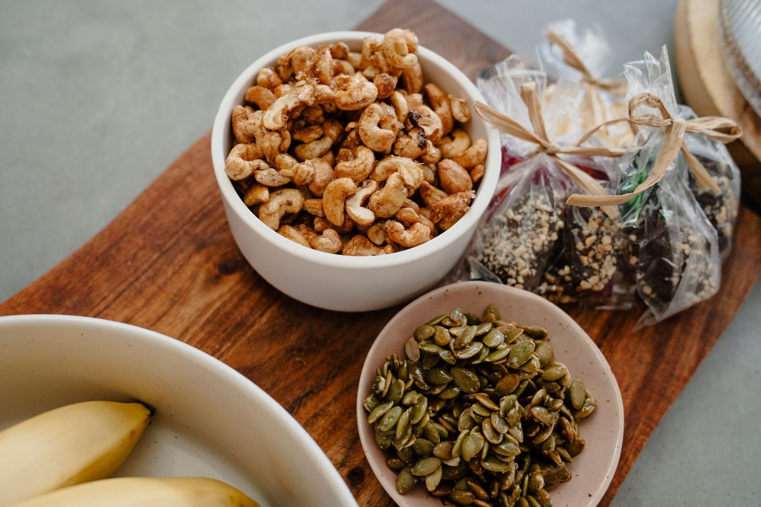 Roasted cashews and pumpkin seeds in white bowls on a wooden board with bananas nearby.