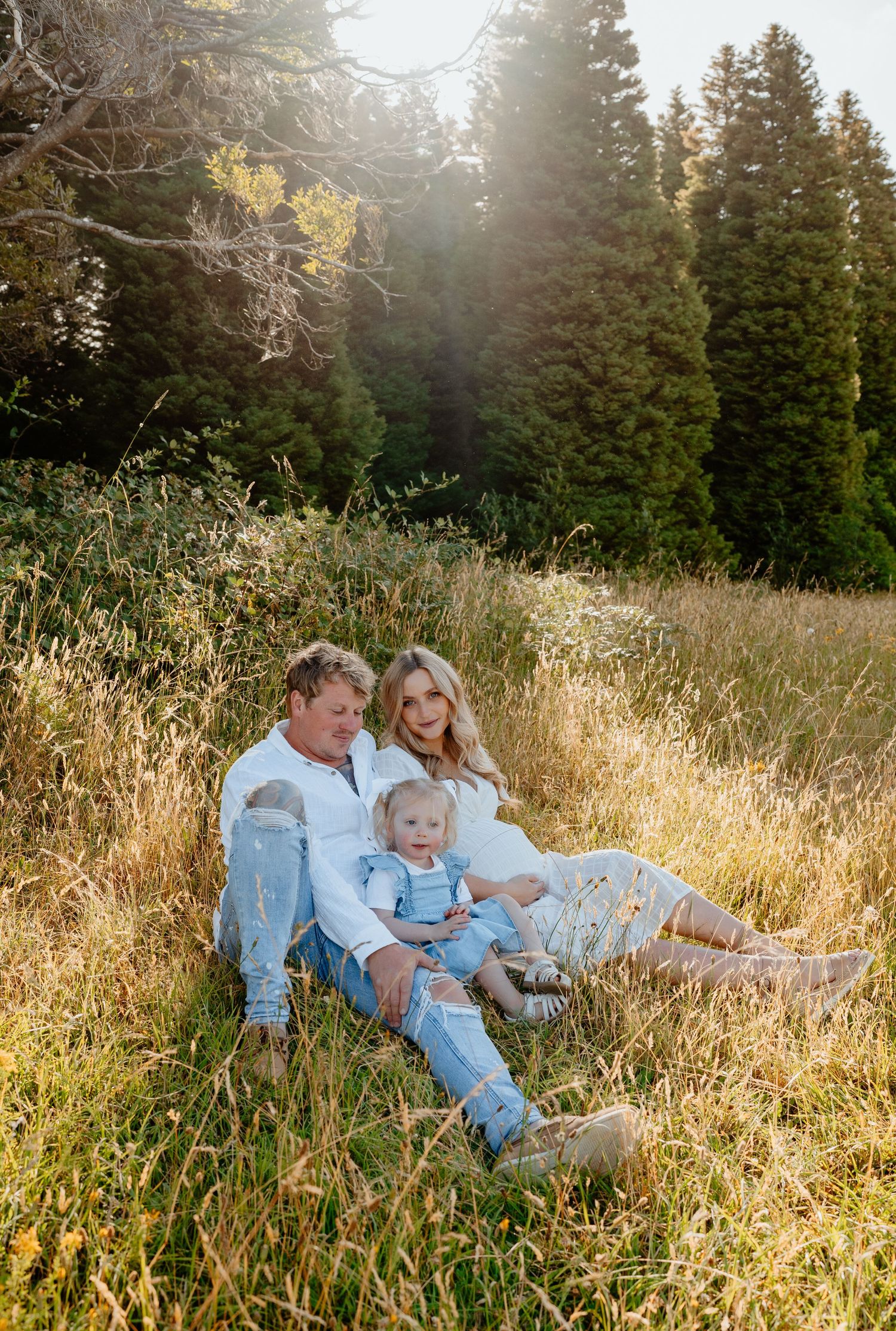 Family of three sitting together in a sunlit meadow surrounded by tall pine trees.