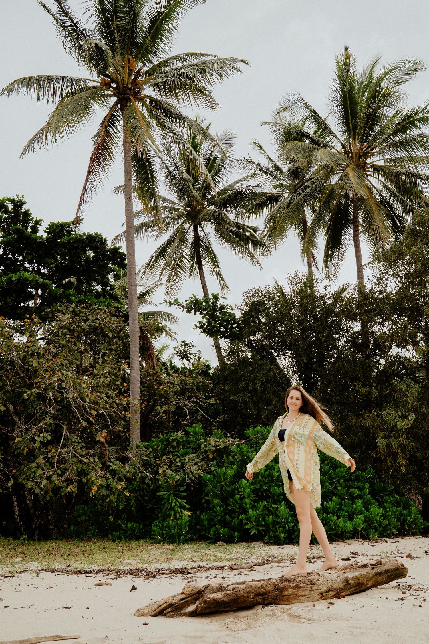 Woman in lace cover-up balancing on driftwood with tall palm trees behind her.
