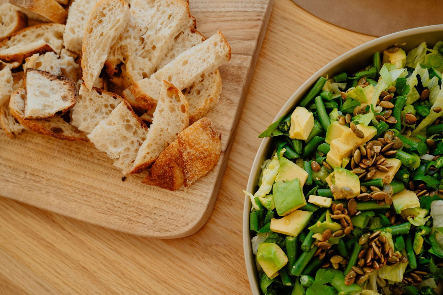 Green salad with avocado, green beans, and seeds served alongside sliced bread on a wooden board.