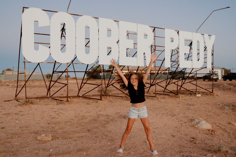 Woman posing with arms raised in front of large white Coober Pedy sign in South Australia.