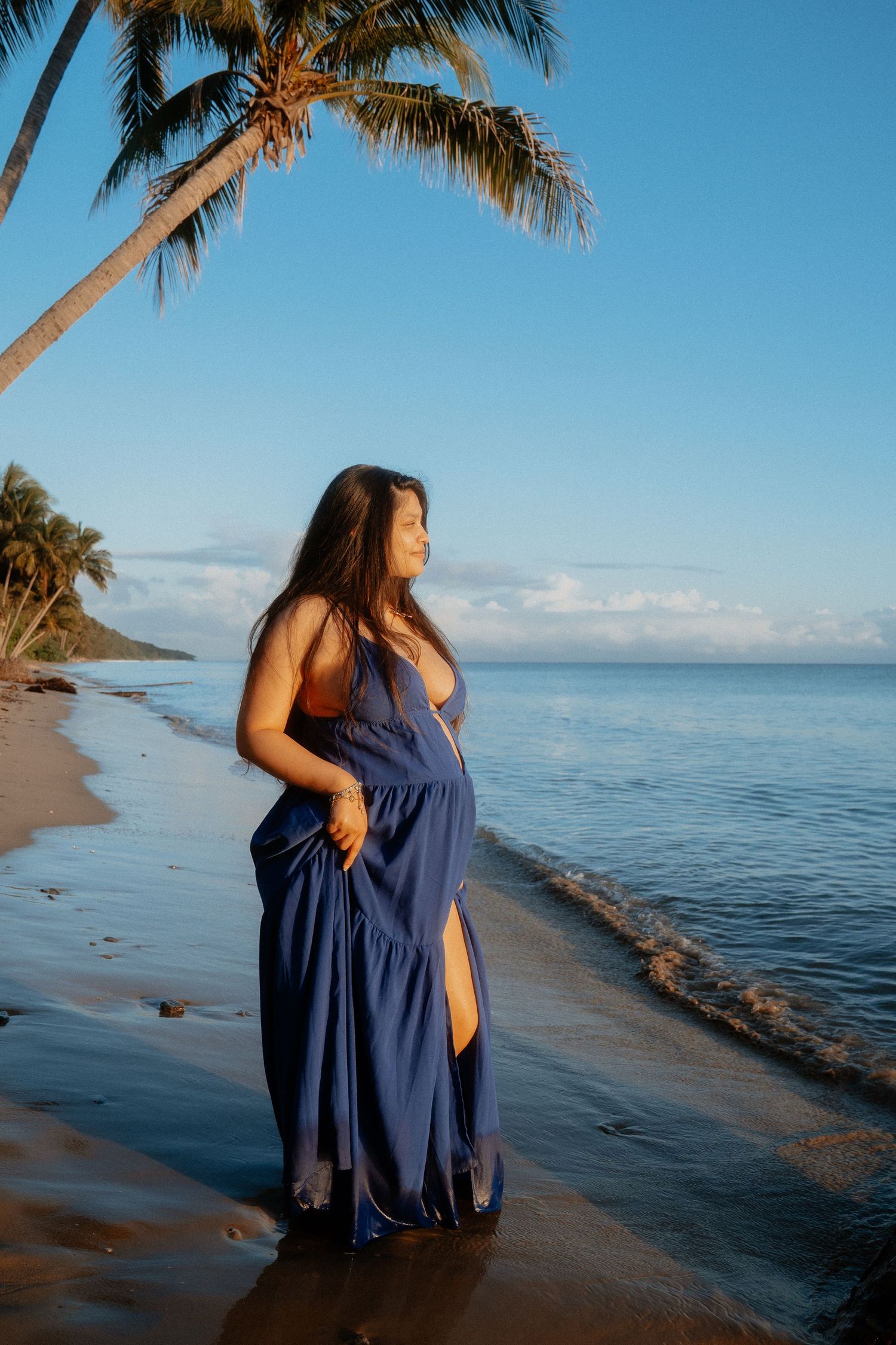 Pregnant woman in blue maxi dress standing on tropical beach at sunset with palm trees.