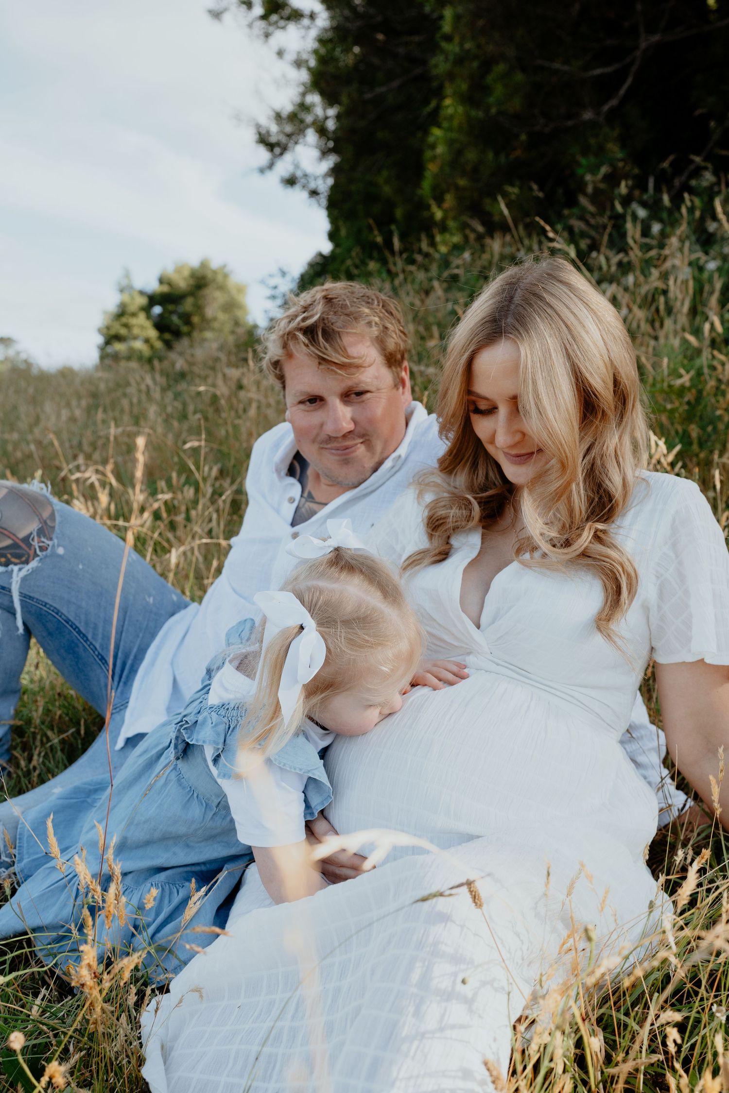 Pregnant woman with husband and toddler daughter relaxing in a golden grass field.