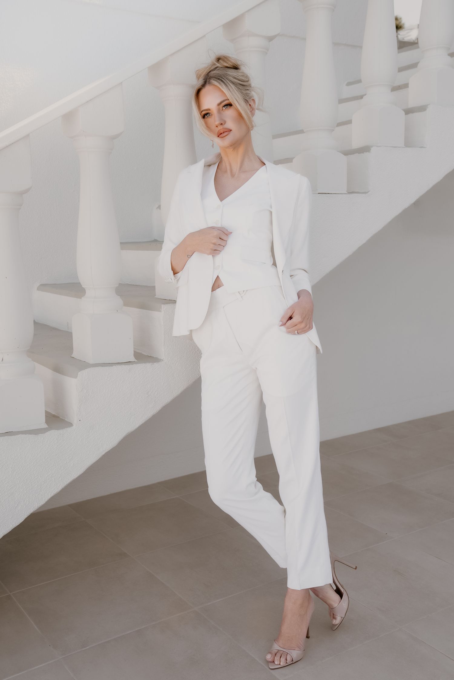 Elegant blonde woman in a white blazer suit posing on a white staircase.