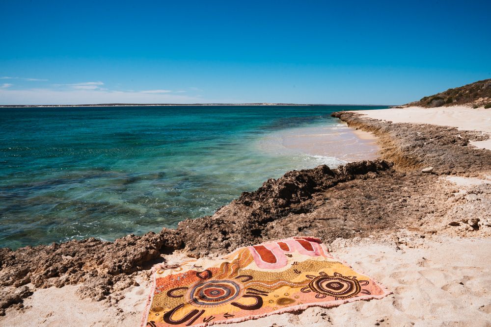 Colorful Aboriginal art print beach towel laid on sandy Australian coastline with turquoise water.