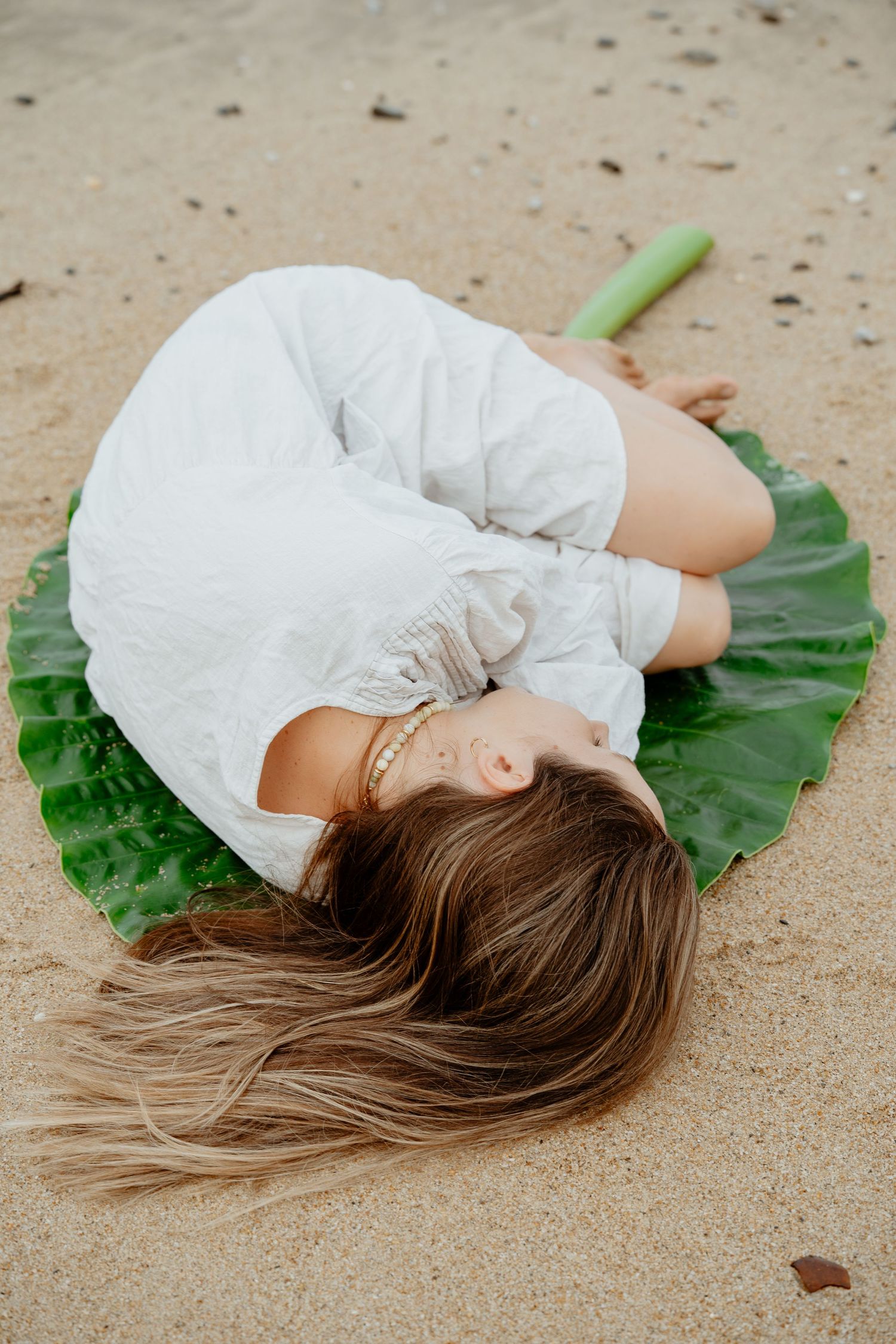 Woman in white dress lying on a large green tropical leaf on sandy beach with eyes closed.
