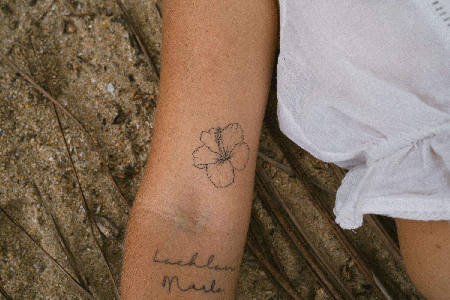 Close-up of a forearm tattoo featuring a hibiscus flower and cursive script names.
