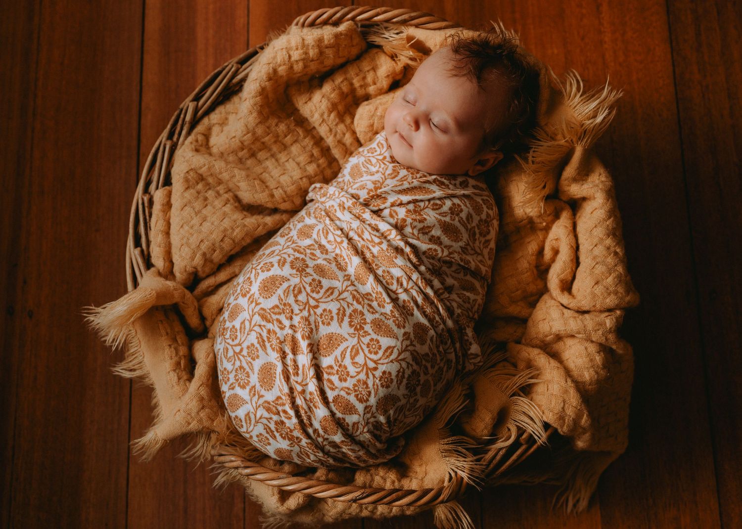 Sleeping newborn baby swaddled in floral wrap nestled in wicker basket with soft tan blanket.