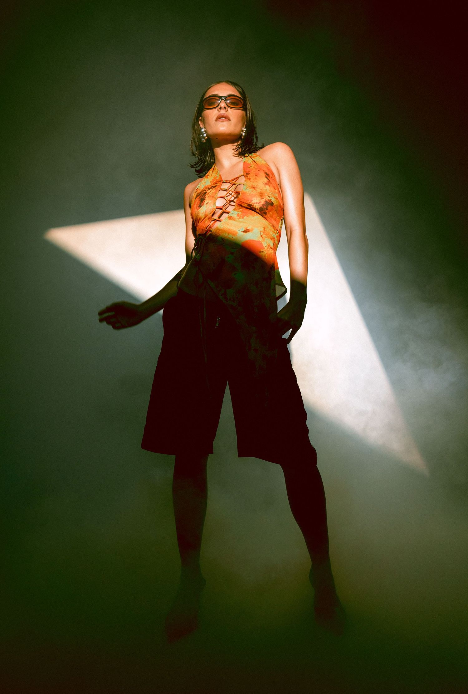 Fashion model in orange tie-dye halter top and black shorts posing in dramatic spotlight beam.
