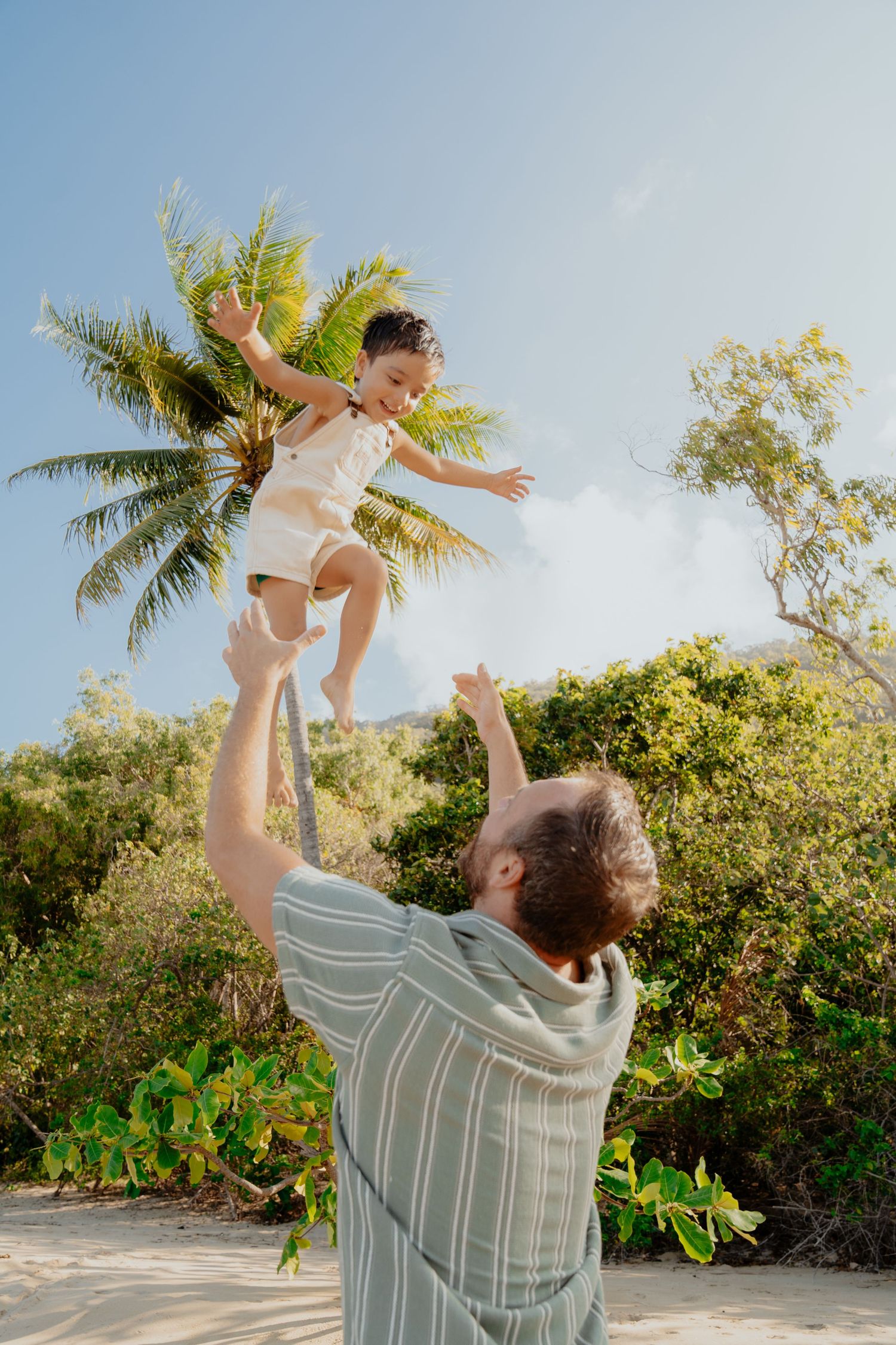 Father joyfully tosses young child in air on tropical beach with palm trees.