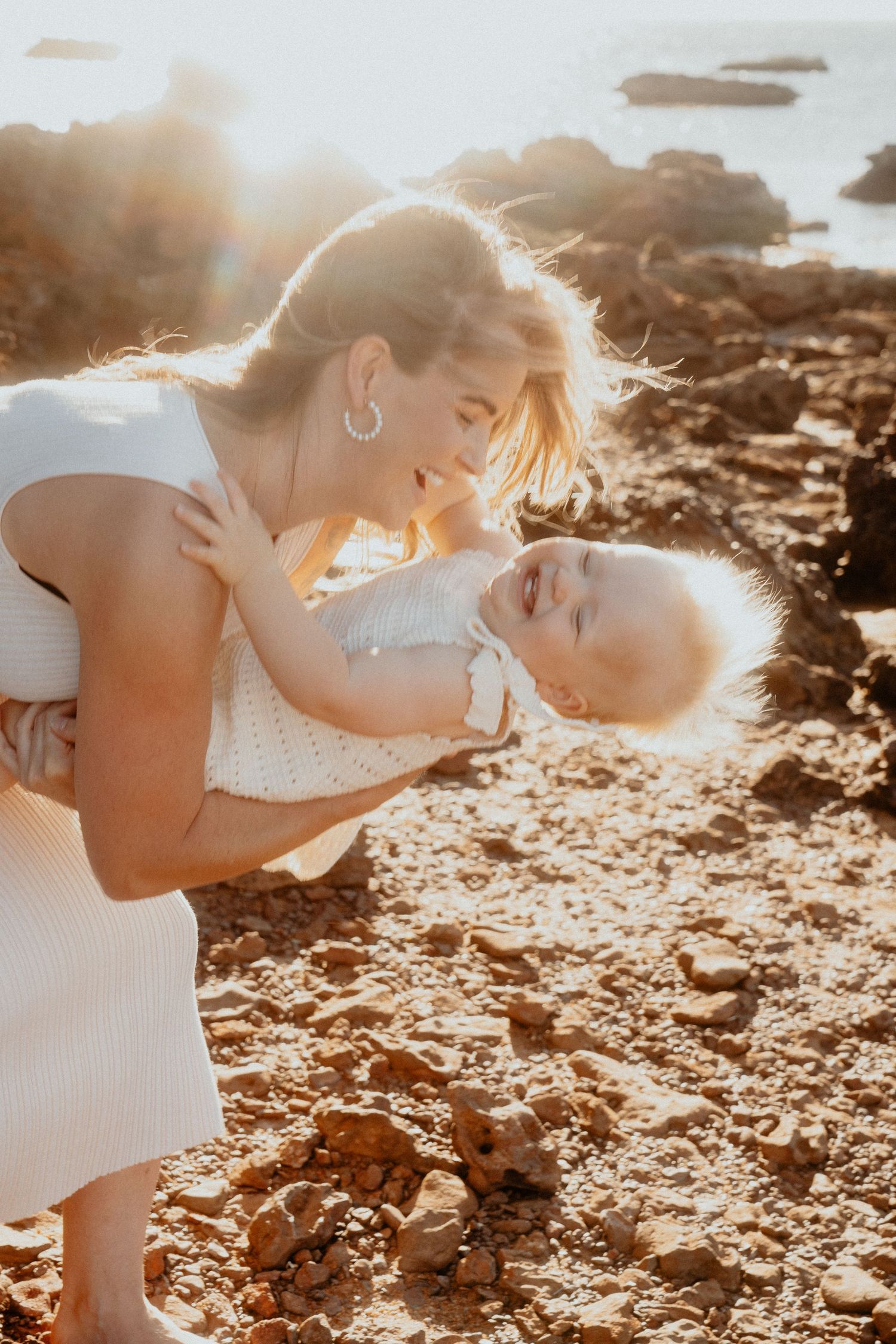 Mother laughing while playfully holding her toddler upside down on a sunny rocky beach at golden hour.