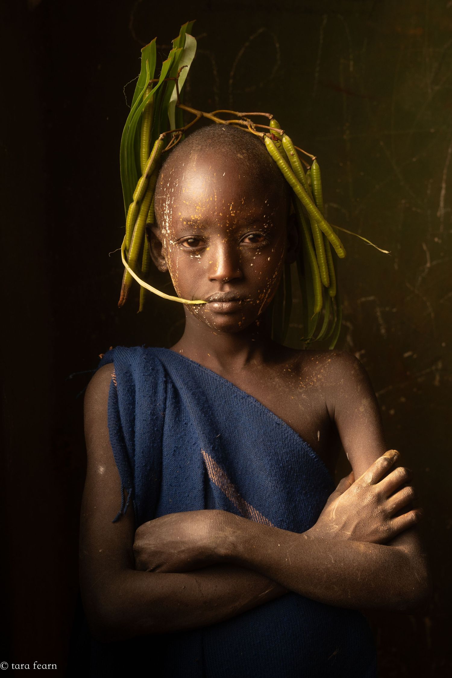Young African child with leaves and gold hoop on head, wearing blue cloth, arms crossed in dramatic portrait lighting.