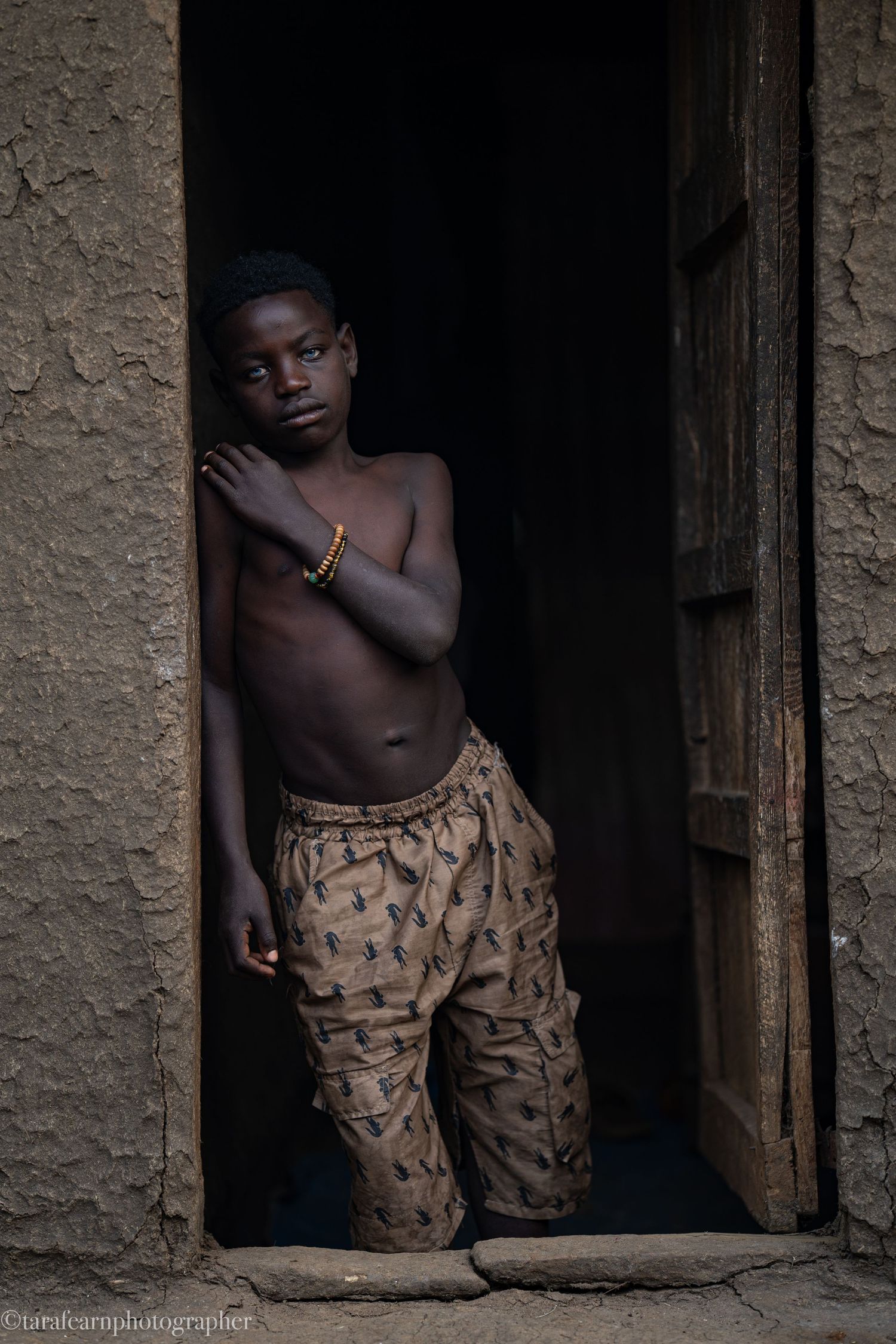 Shirtless African boy wearing patterned pants and a gold bracelet standing in a rustic wooden doorway.