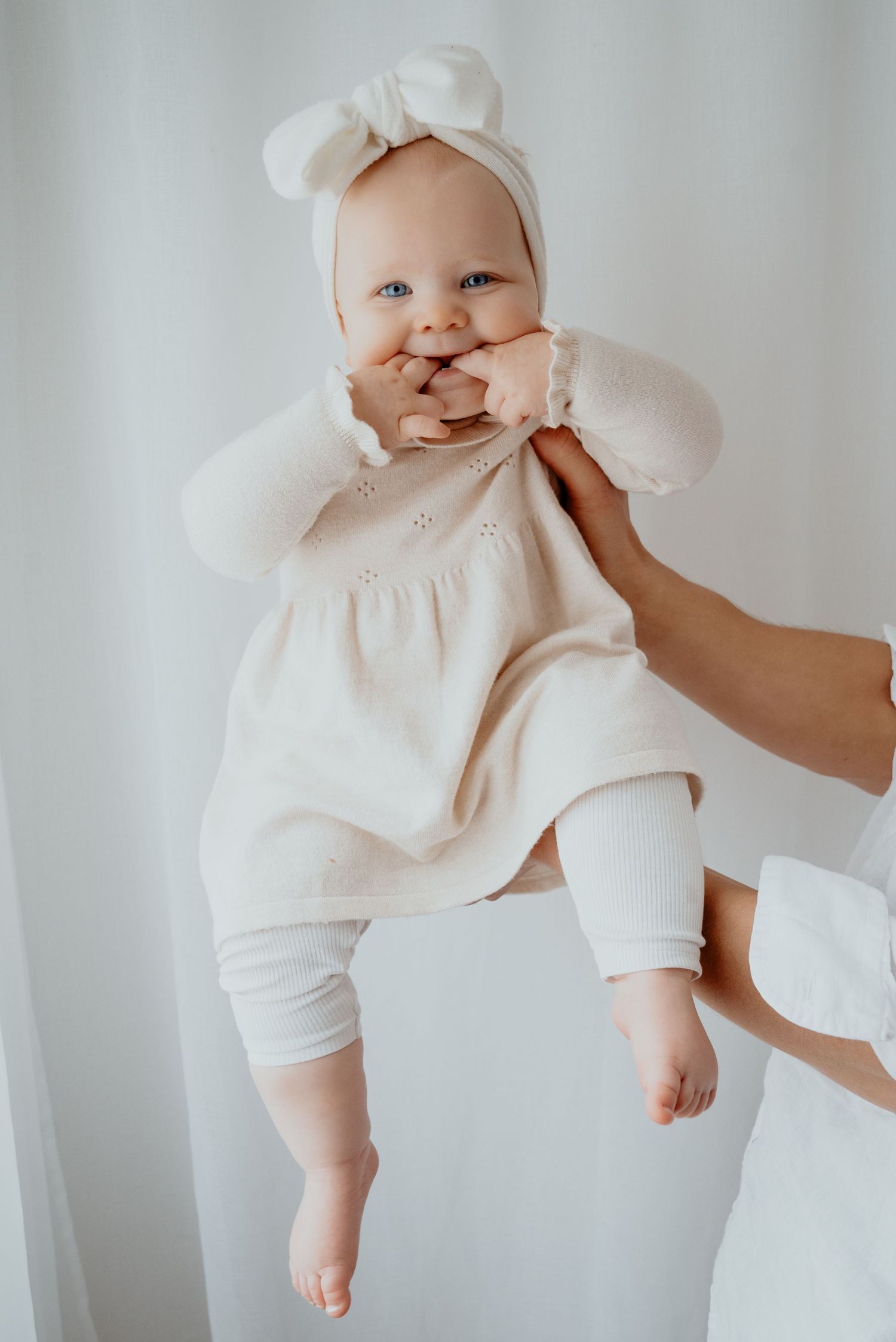 A smiling baby in a cream dress and bow headband is held up by adult hands against a soft white background.