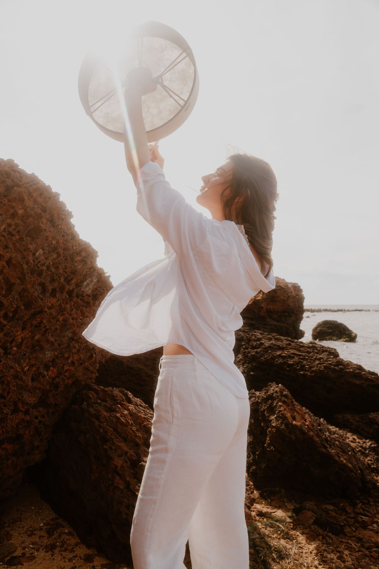 Woman in white outfit holding a hat up against sunlit rocky coastal scenery.