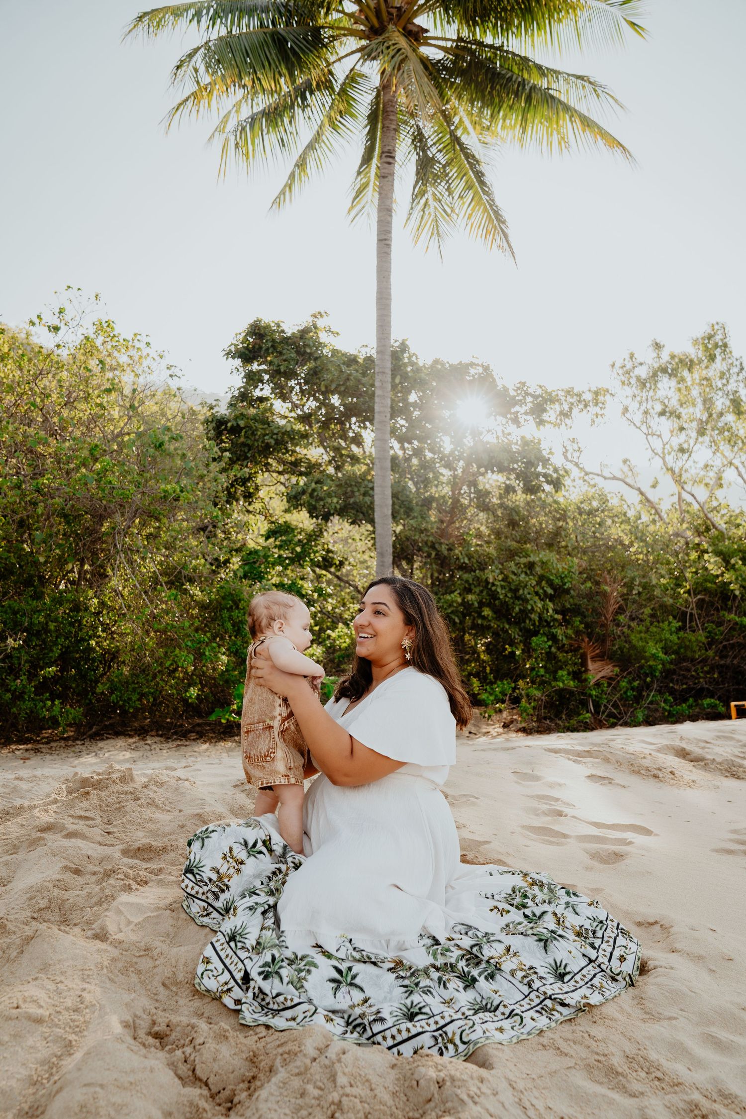 A smiling woman in a white floral dress sits on sandy beach holding a baby under a tropical palm tree at sunset.