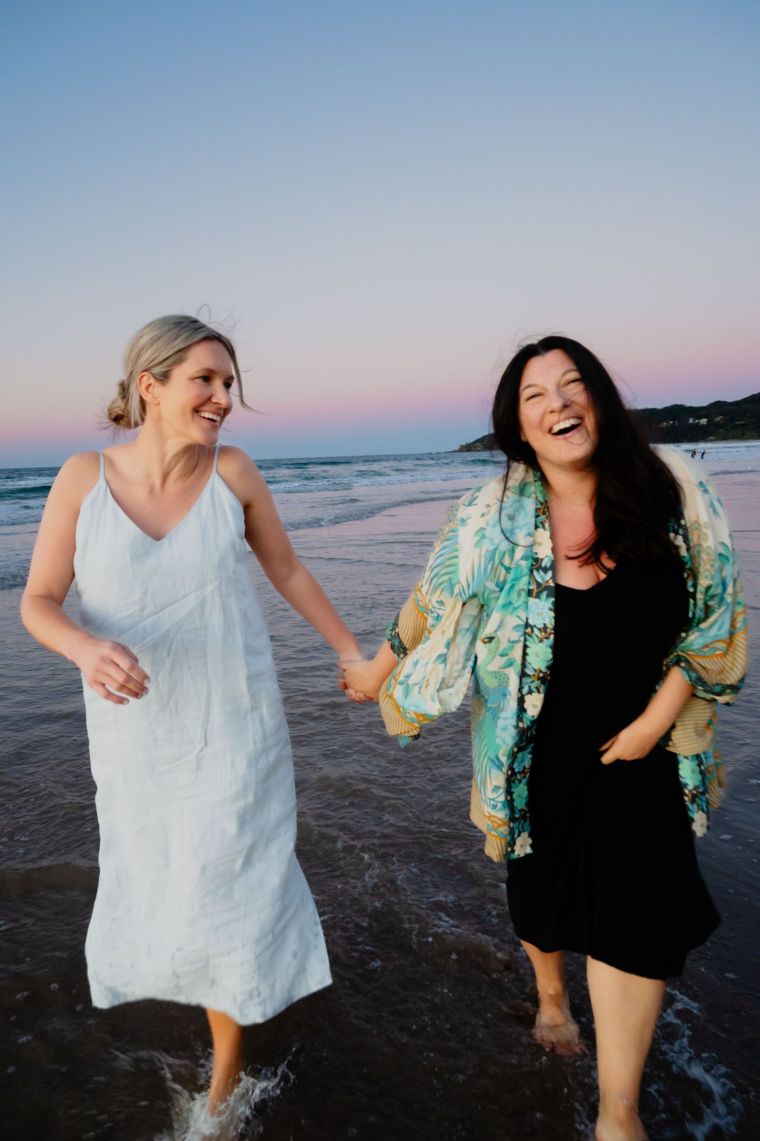 Two women holding hands and laughing while wading through ocean waves at sunset.