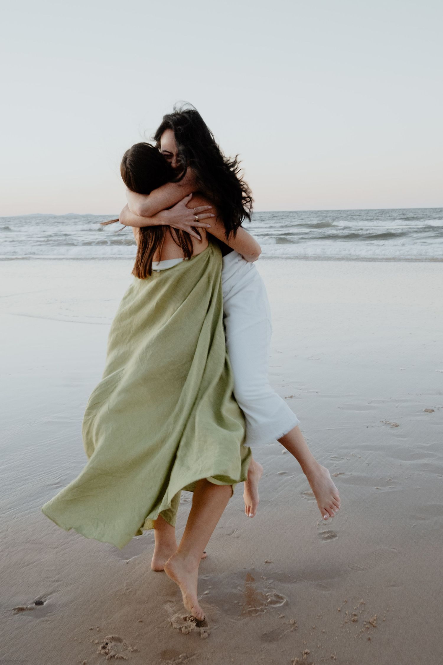Two women embracing joyfully on a sandy beach at dusk, one wearing a green dress.